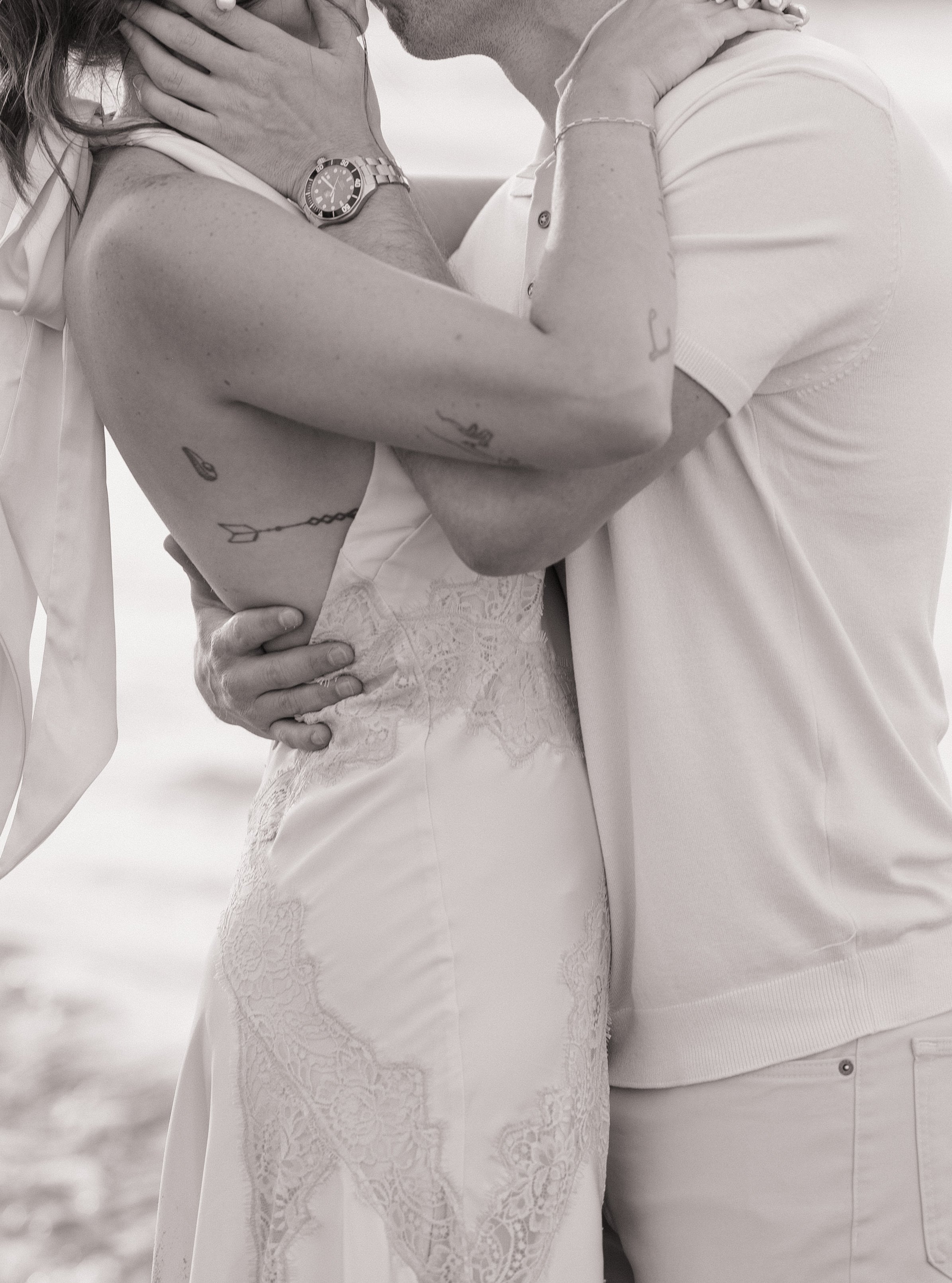 A couple sharing an intimate moment, close-up of their torsos and arms, with the woman in a lace dress and the man in a white polo, both holding each other gently.
