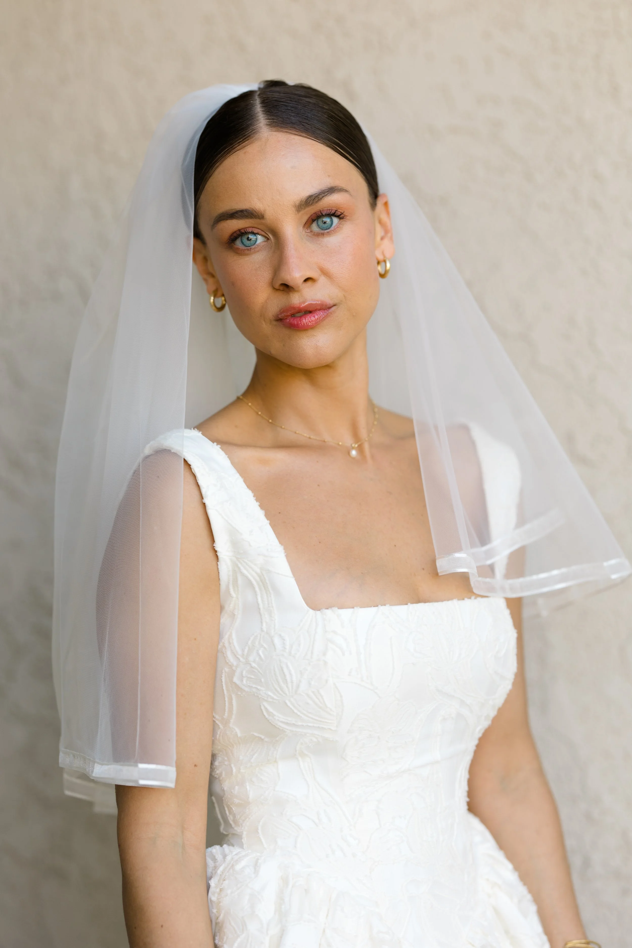 A woman with blue eyes, dark hair in a sleek style, wearing a white wedding dress with embroidered details, gold hoop earrings, a delicate necklace, and a sheer veil, standing against a textured beige wall.