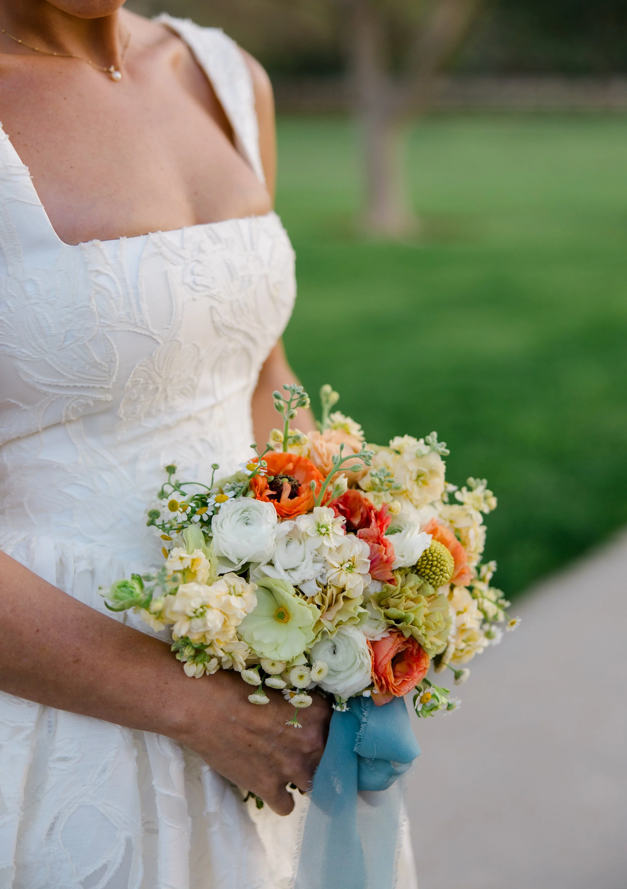 Close-up of a bride holding a colorful bouquet of flowers, wearing a white embroidered wedding dress with a pearl necklace, outdoors with blurred greenery in the background.