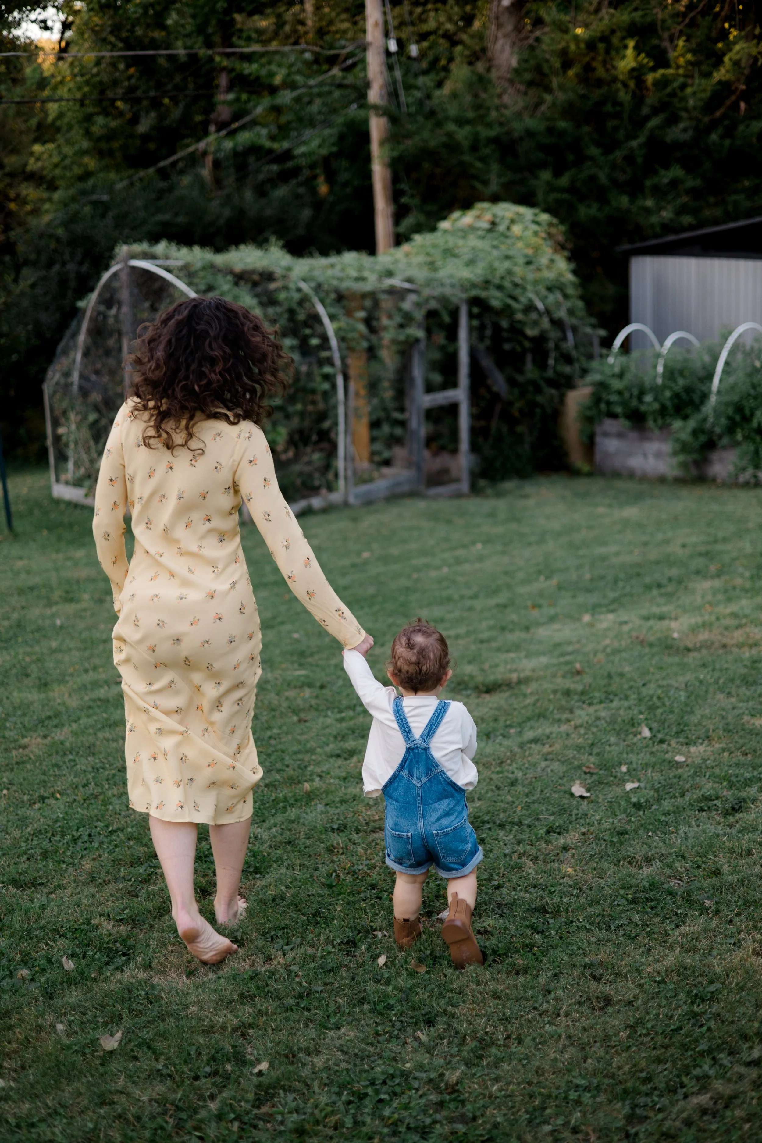 A woman in a yellow floral dress walking hand-in-hand with a young child in denim overalls on a grassy lawn, with a backyard garden and trees in the background.