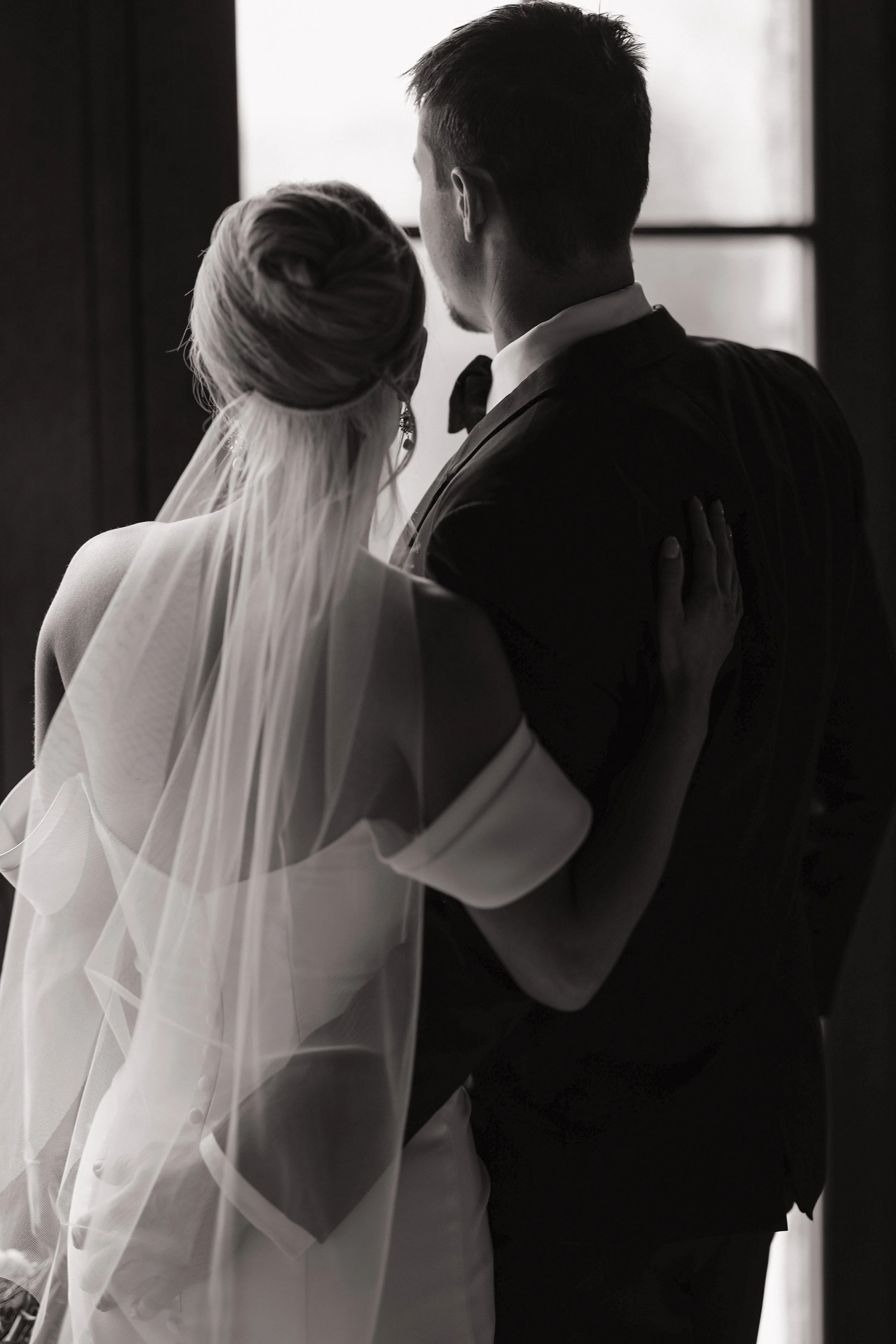 Black and white photo of a bride and groom standing close together indoors, facing away from the camera, looking out a window.