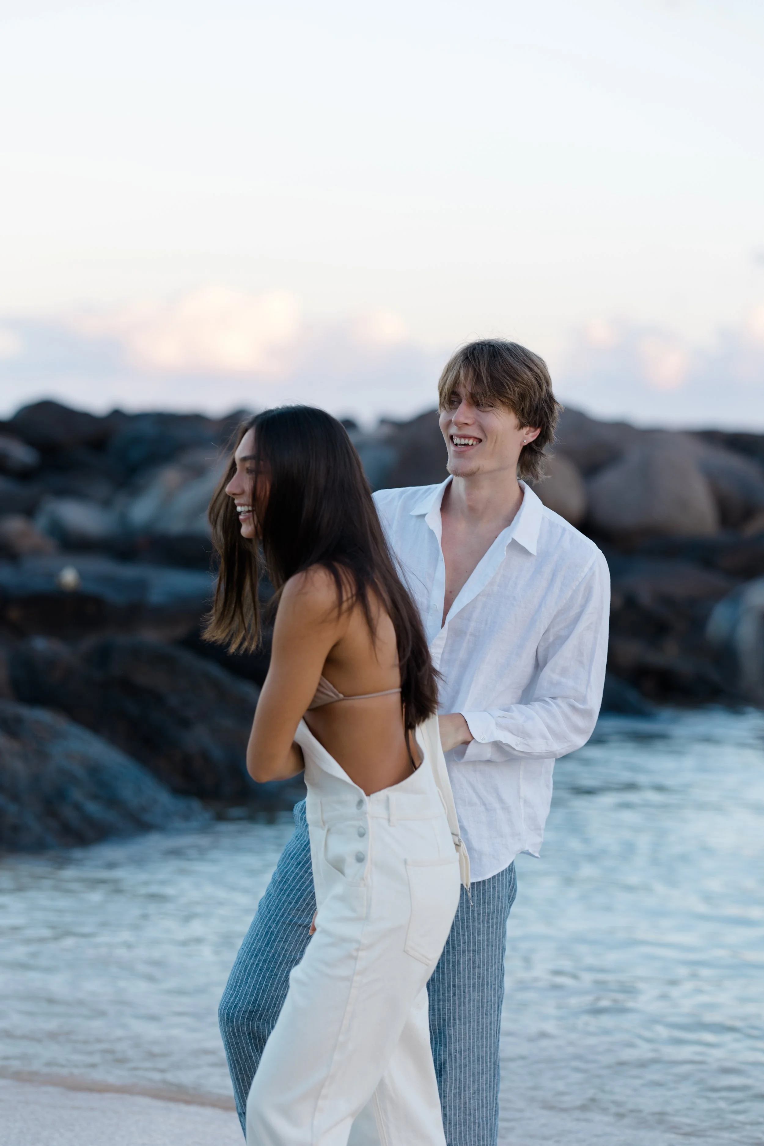A young man and woman are standing on a rocky beach with the ocean and sky in the background, smiling and enjoying the moment.