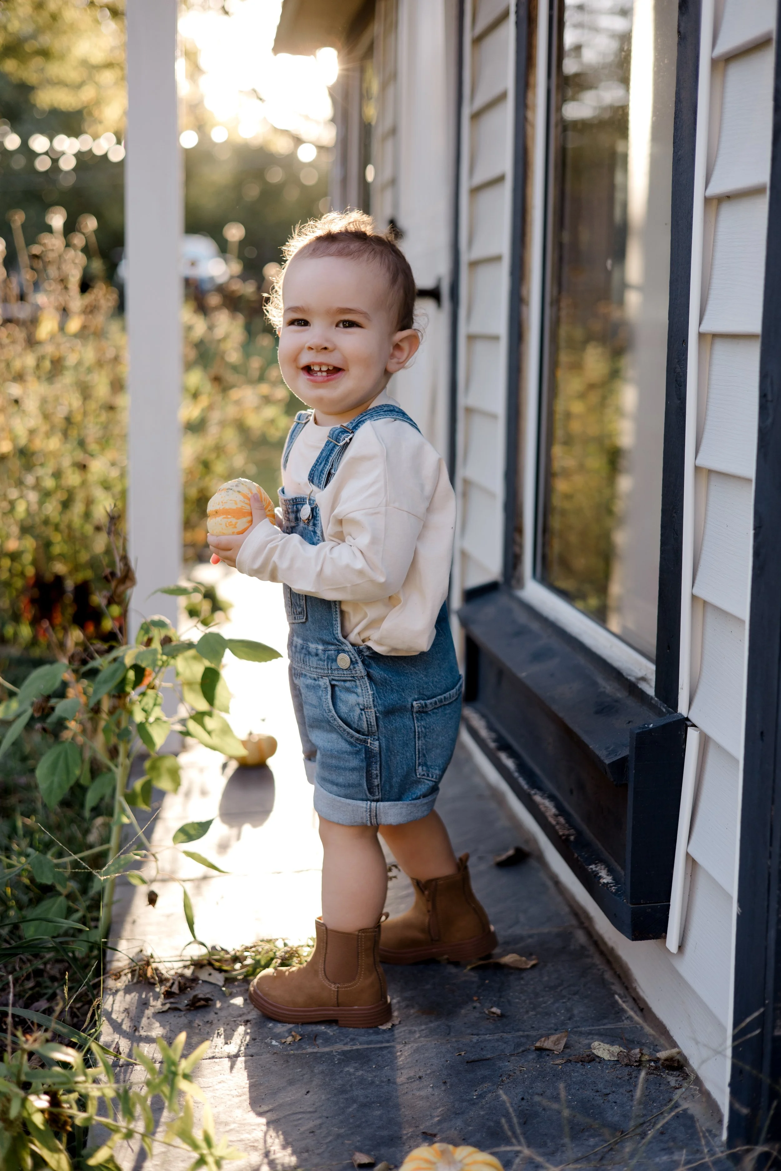 A young boy with short brown hair, wearing denim overalls, a cream-colored long-sleeve shirt, and brown boots, standing on a porch near a garden, holding a small pumpkin, smiling at the camera during sunset.