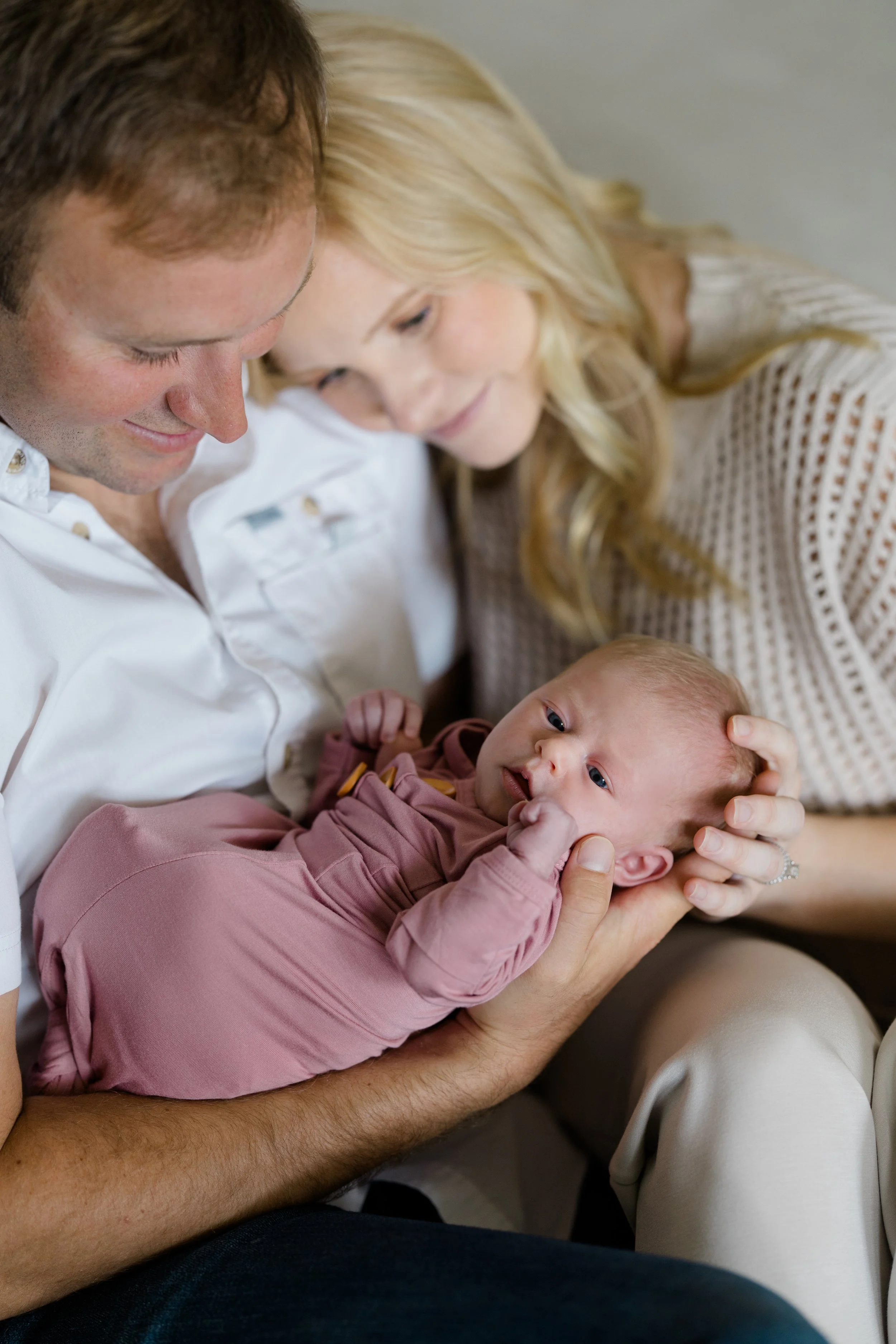 A man and woman lying close together, smiling and looking down at a baby, who is being held in the man's arms.