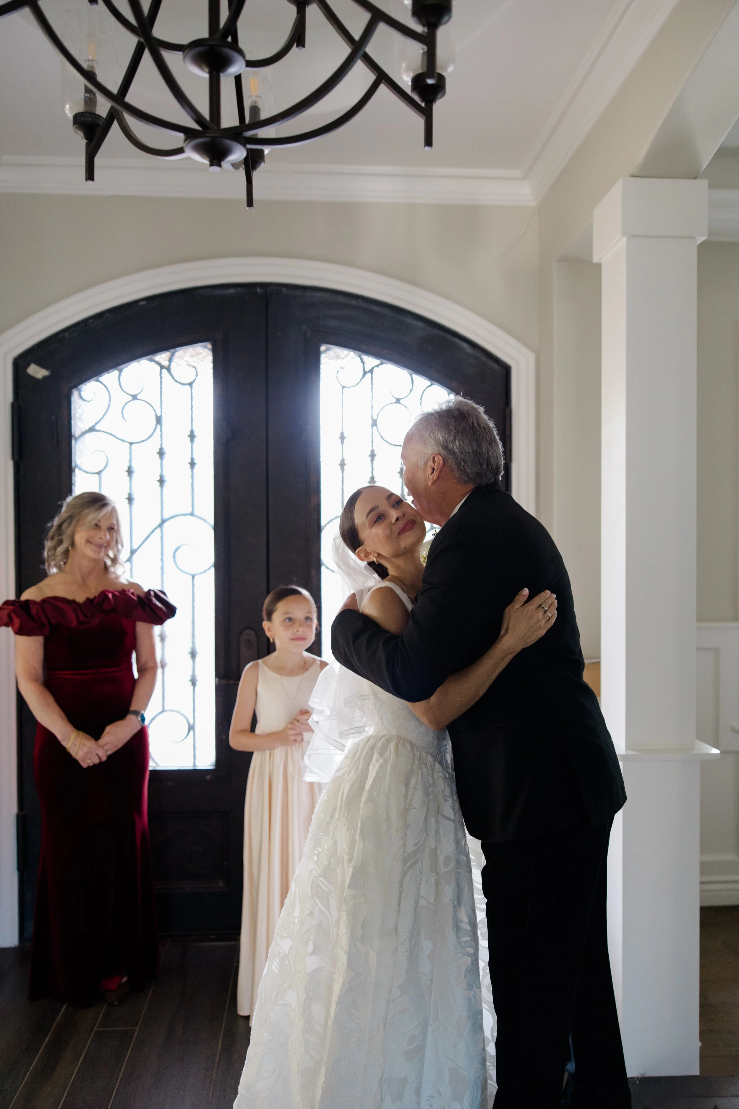 A bride and groom are sharing a dance in a wedding ceremony. An older woman in a maroon dress and a young girl in a beige dress are watching nearby. The scene takes place inside a house with a large front door and a chandelier overhead.