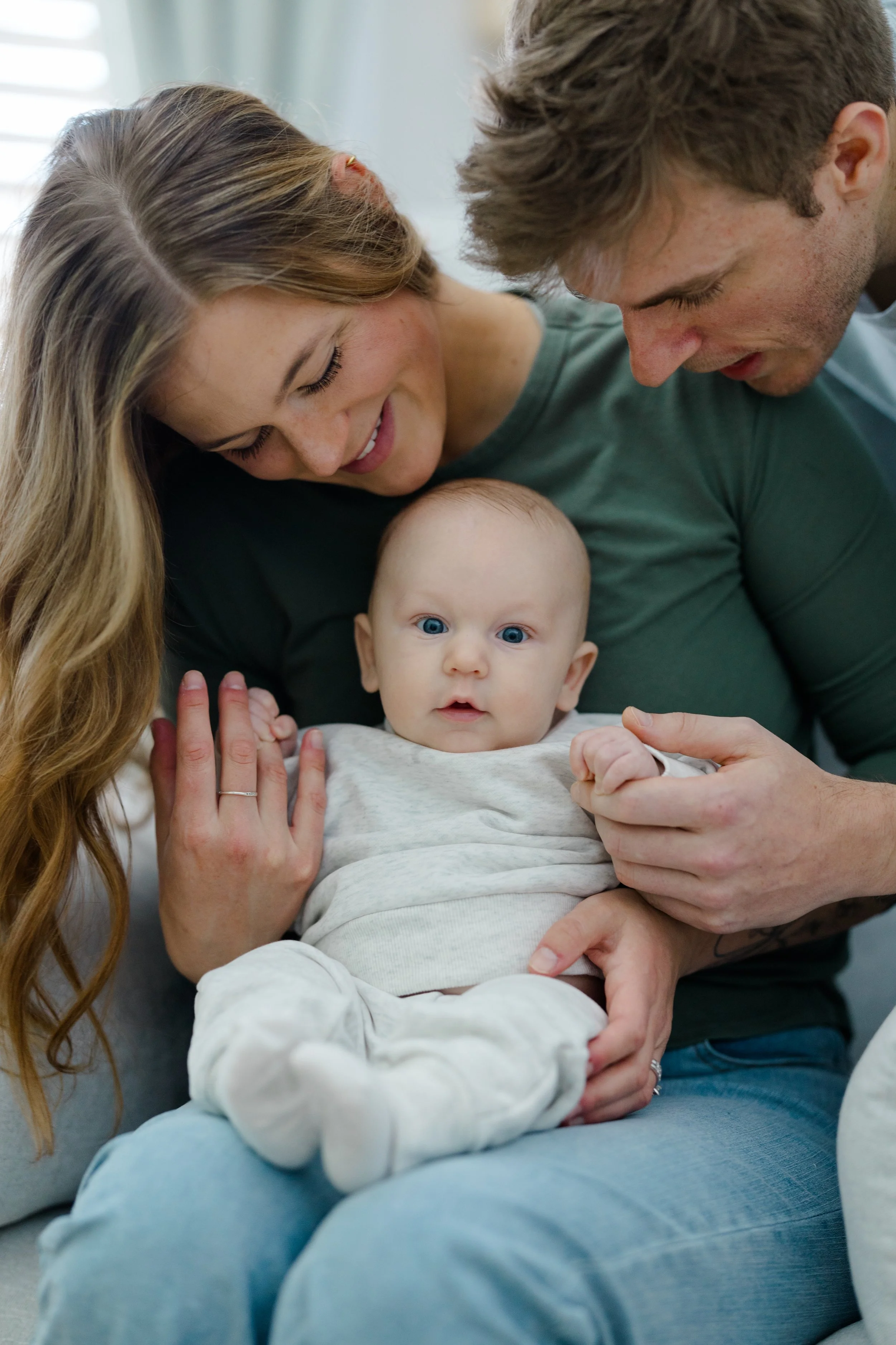 A family of three enjoying a close moment together, with the mother and father leaning over their baby who is lying on her back, looking up.