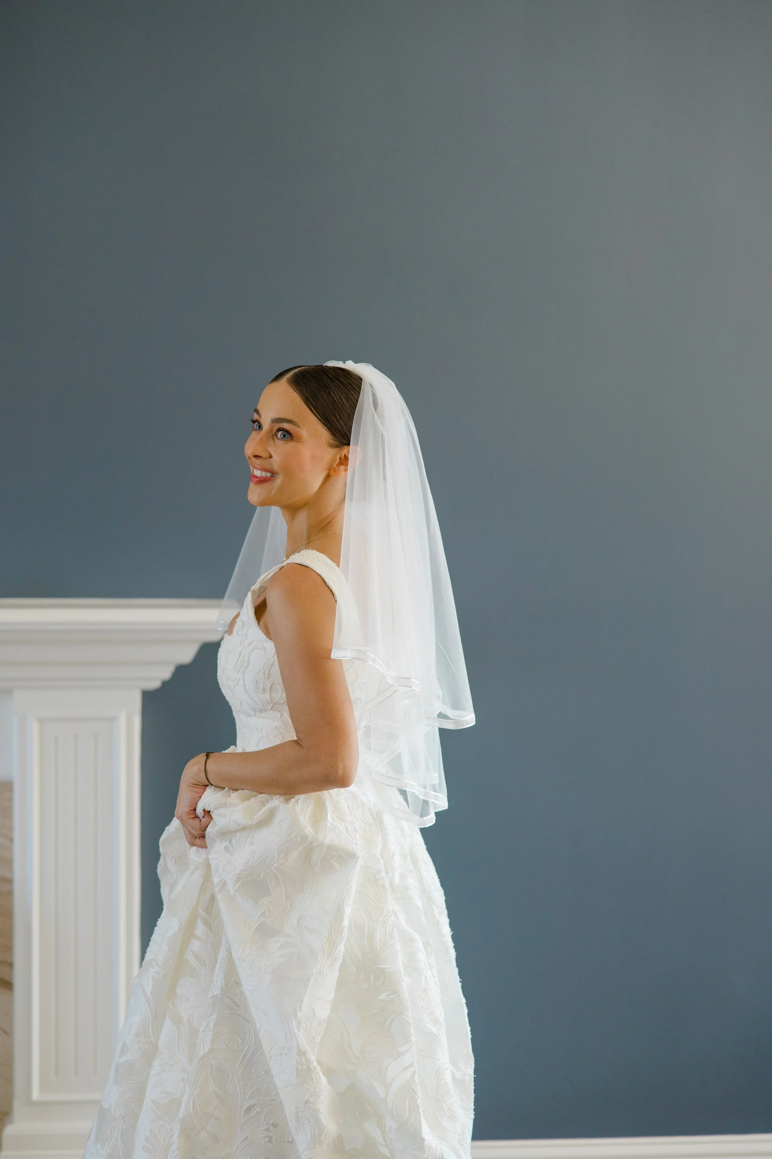 A woman in a white wedding dress and veil standing indoors with a blue-gray wall background and white fireplace mantel.