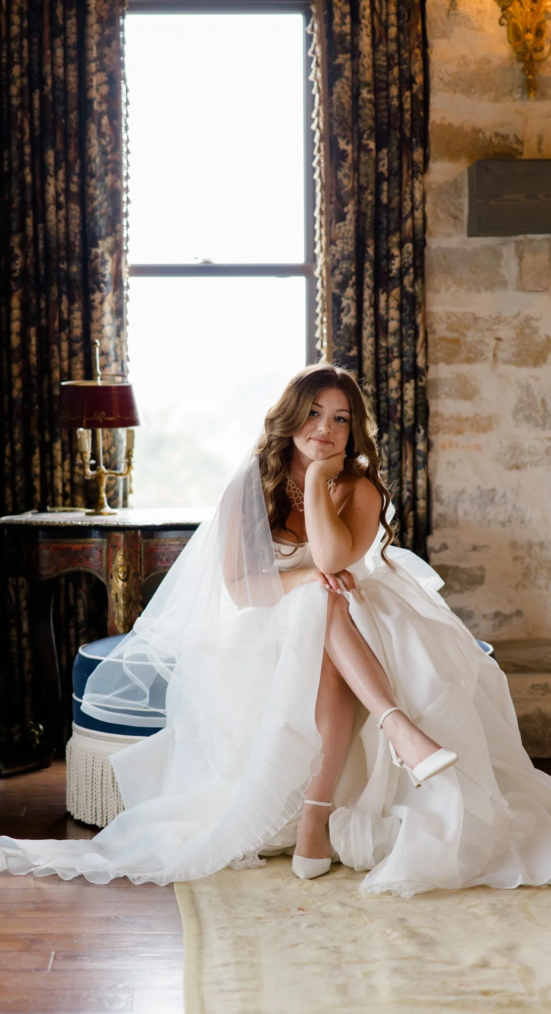 A woman in a wedding dress sitting on a chair, with one leg across the other, in a room with large window and patterned curtains