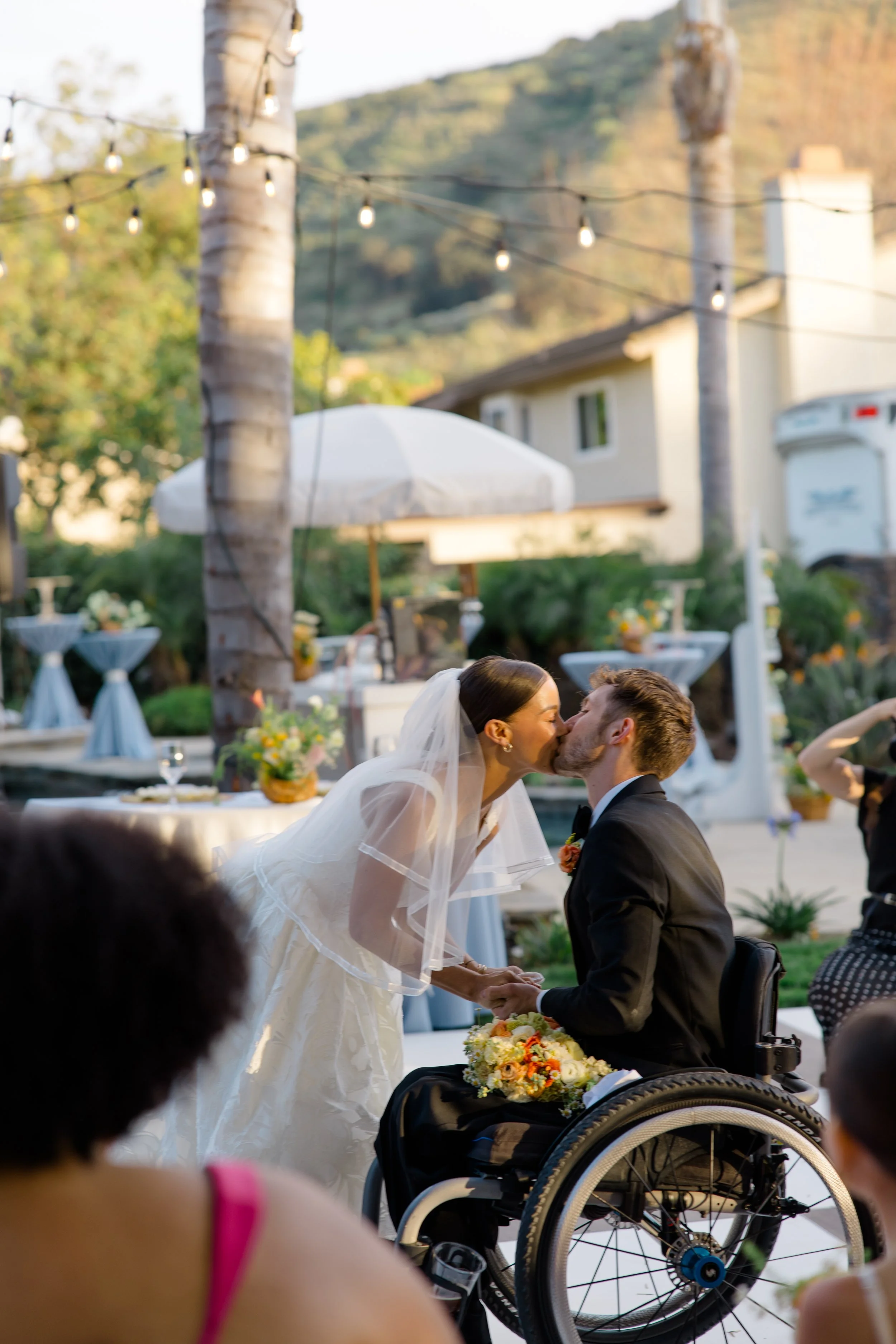 A bride and groom kiss during their wedding ceremony outdoors, with guests watching. The bride is wearing a white wedding dress and veil, and the groom is in a black suit. There are tables with flowers and a white canopy in the background.