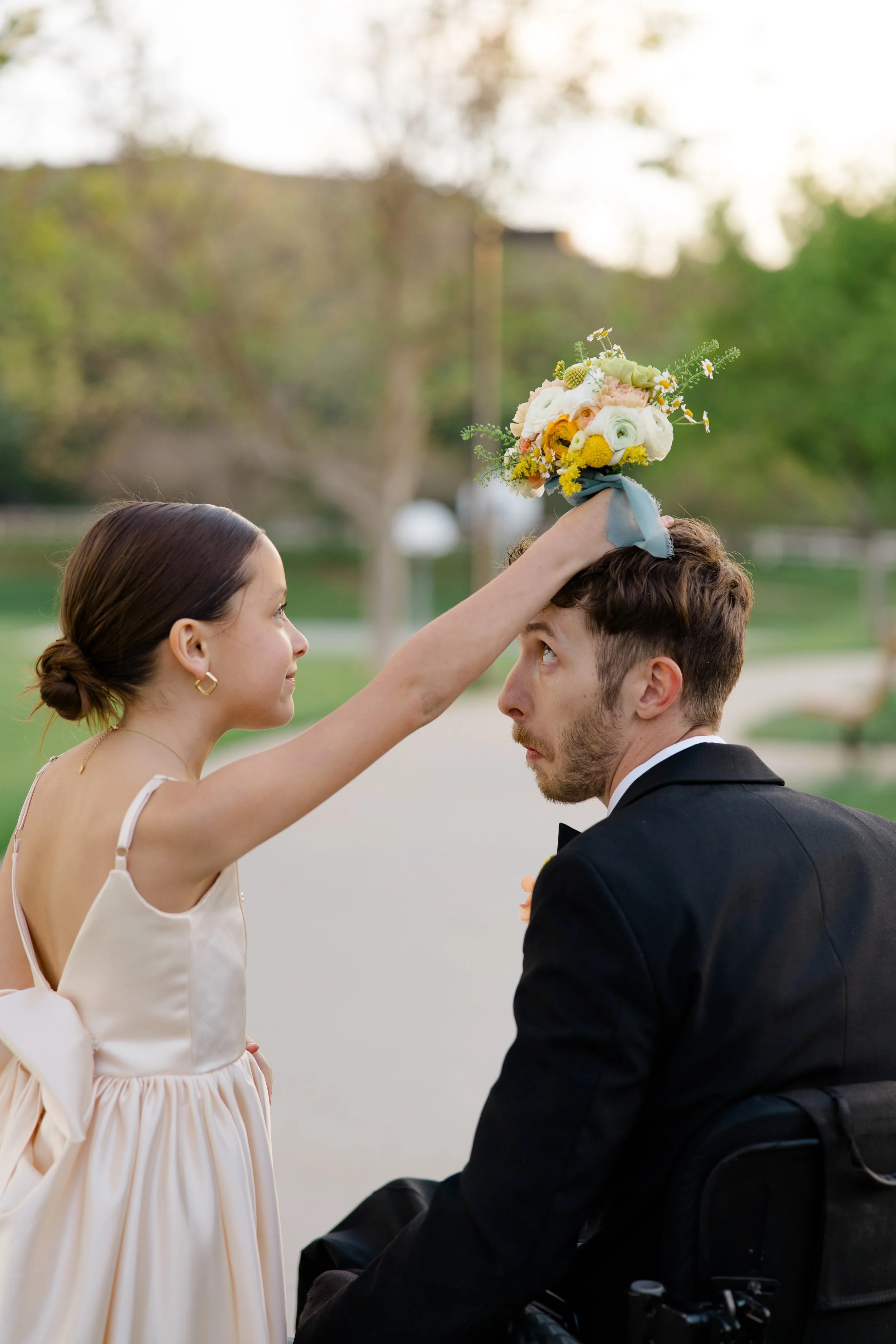 Woman in light-colored dress placing a flower bouquet on a young man's head, who is sitting in a wheelchair, outdoors with blurred trees and sky in the background.