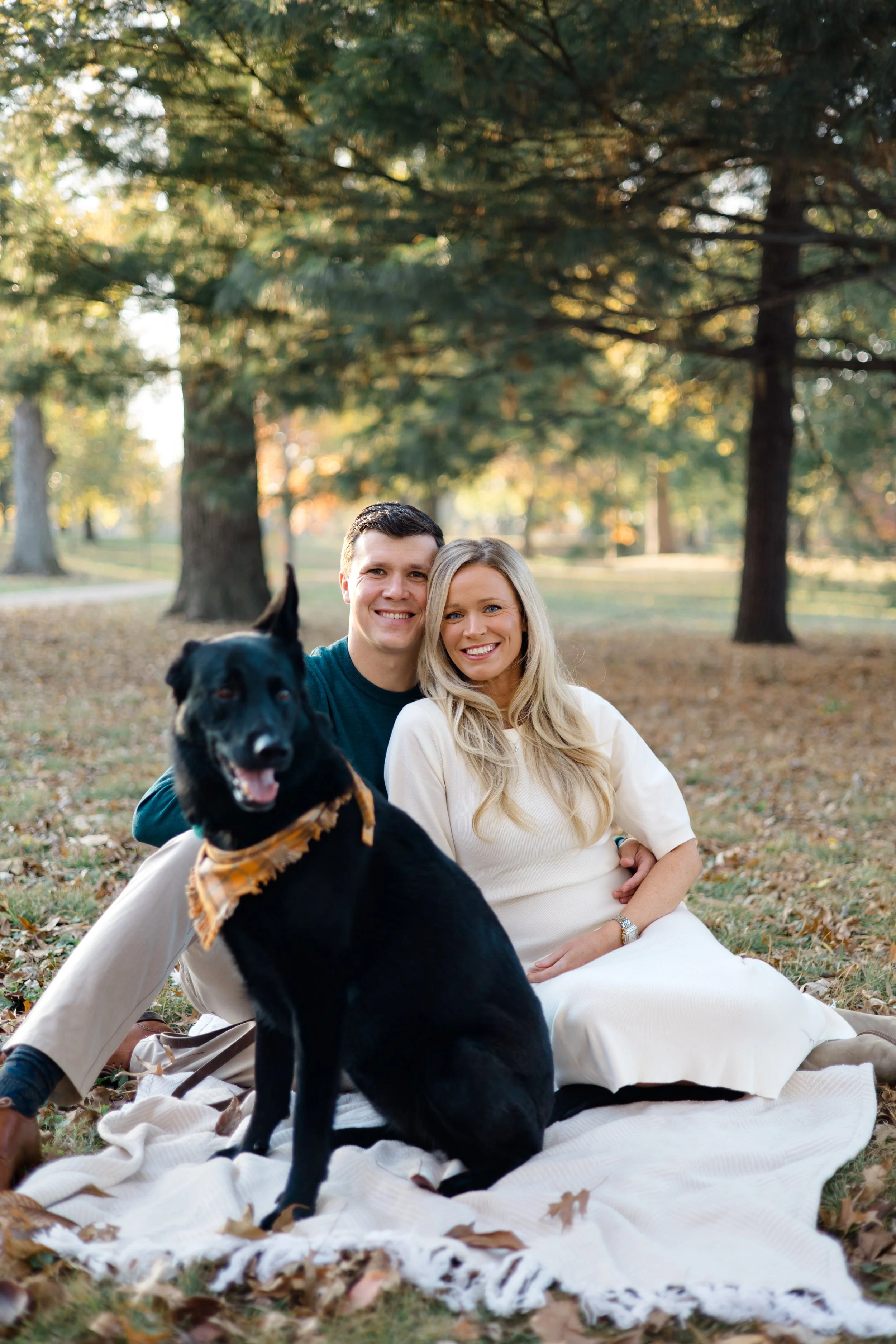 A couple and their black dog sitting on a blanket in a park surrounded by fall leaves and trees with sunlight filtering through the branches.