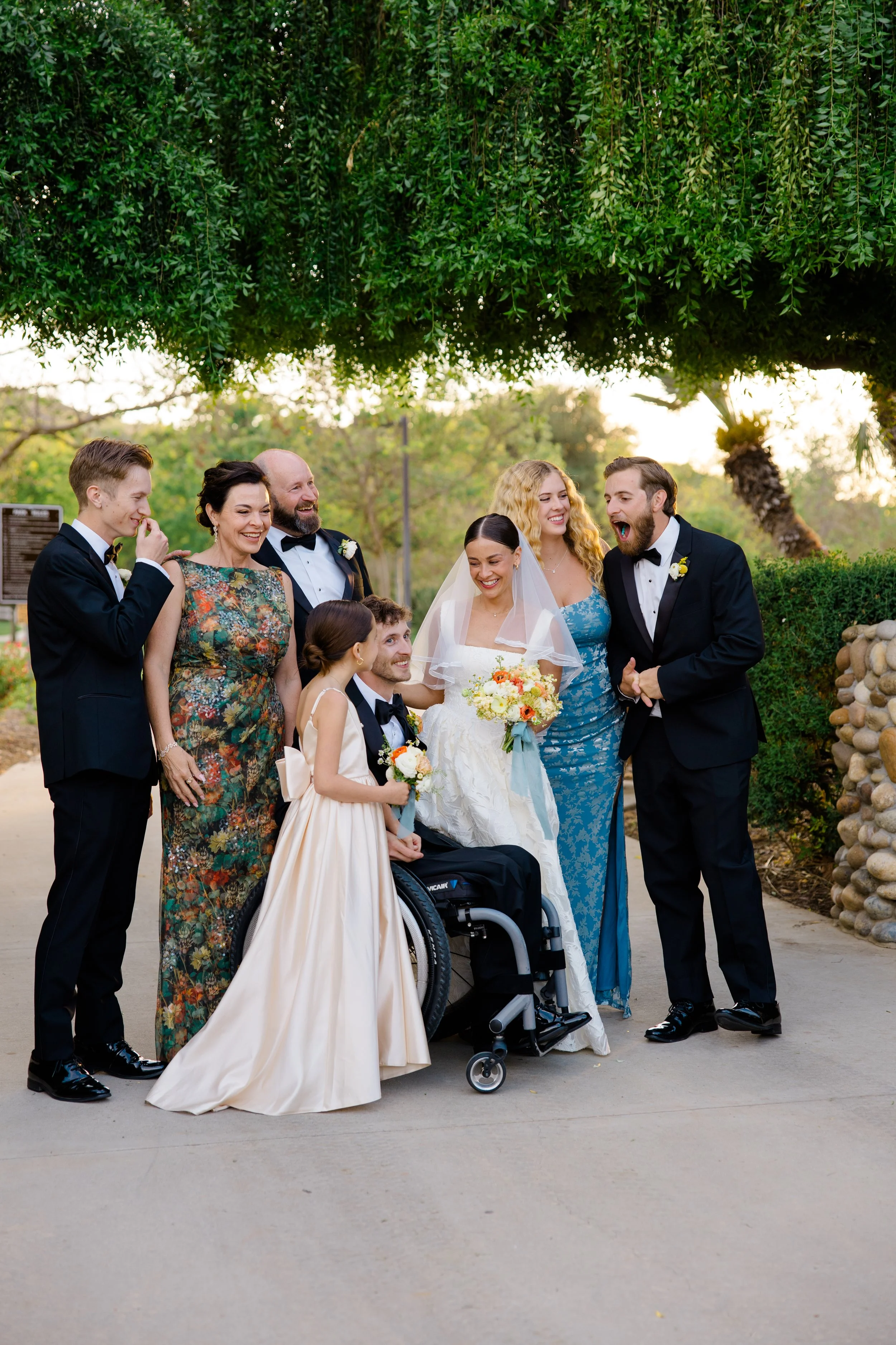 A group of people at a wedding outdoors, including a bride in a white dress and veil, a groom in a tuxedo, and several guests smiling and laughing, some in formal attire, with trees and greenery in the background.