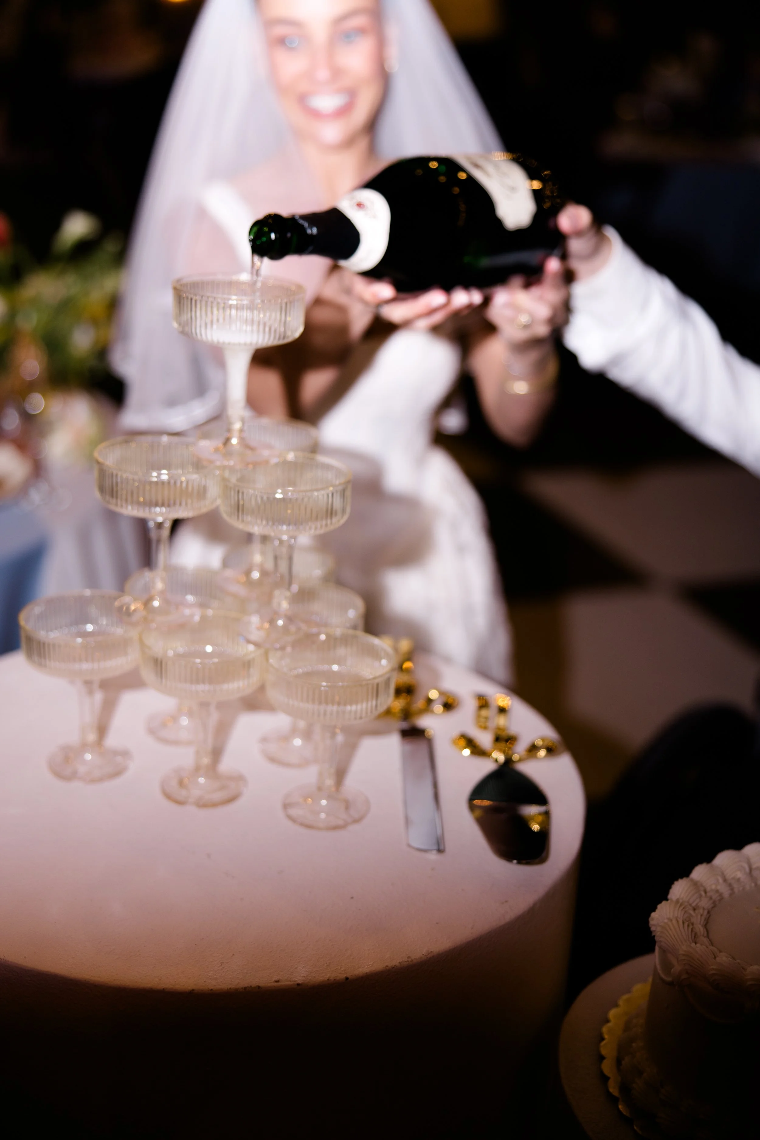 A woman in a wedding dress and veil is smiling and holding a bottle of champagne, pouring it into a tower of glasses at a celebration or wedding event.