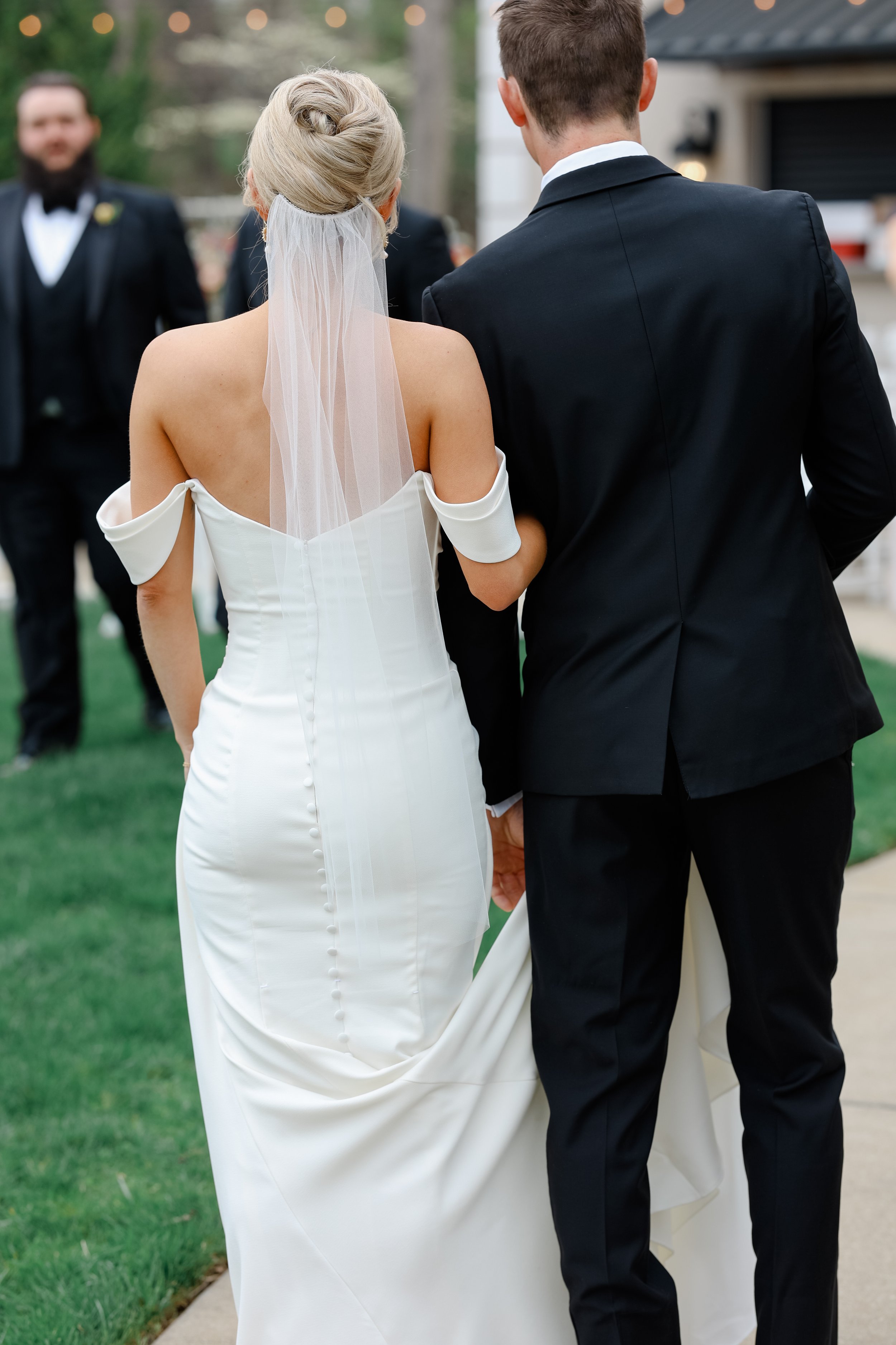 A bride and groom holding hands outside during a wedding ceremony, with the bride in a white off-the-shoulder wedding dress and veil, and the groom in a black suit, with friends or family in formal attire in the background.