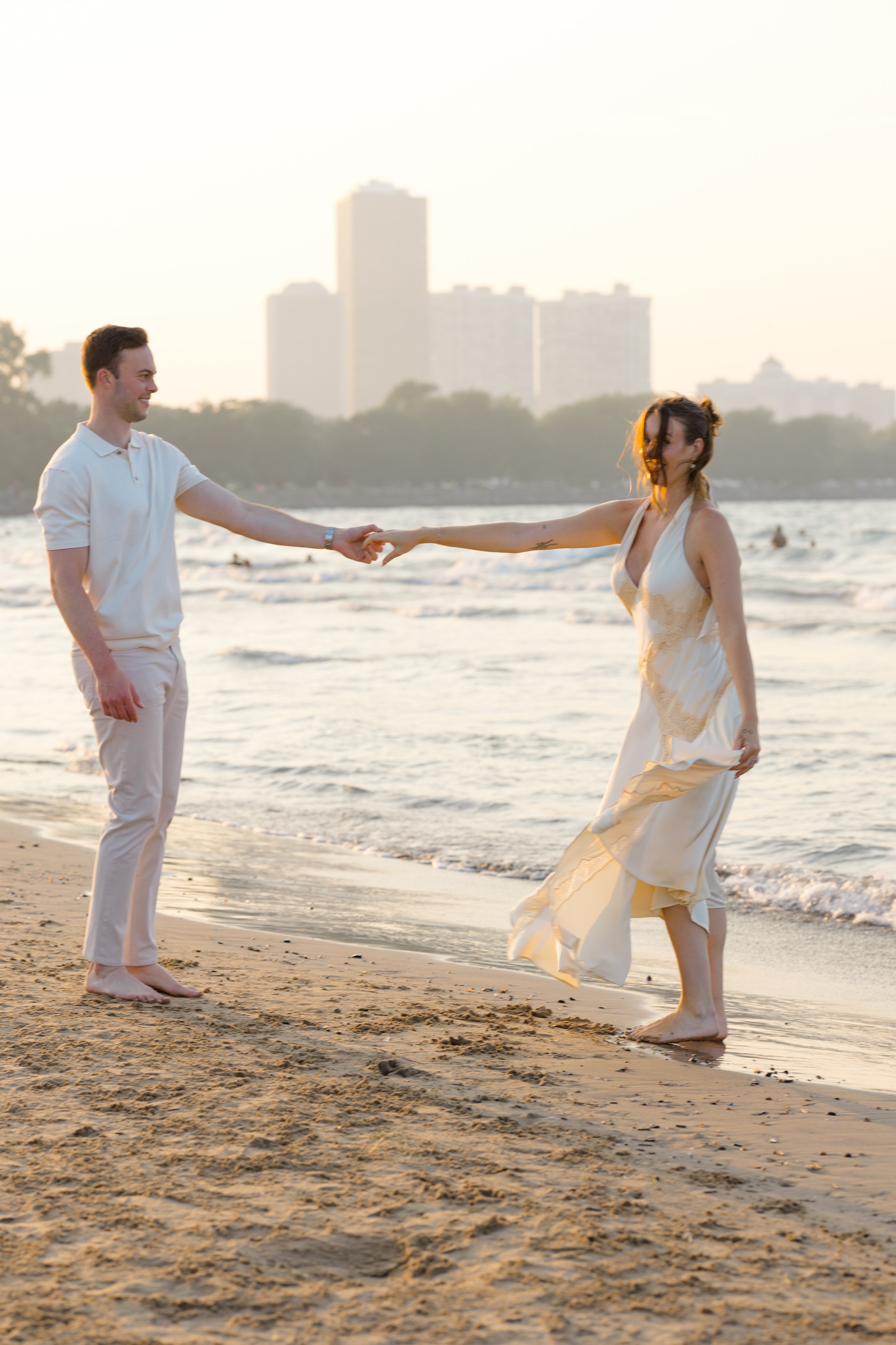 A man and woman holding hands on the beach during sunset, with city buildings in the background.
