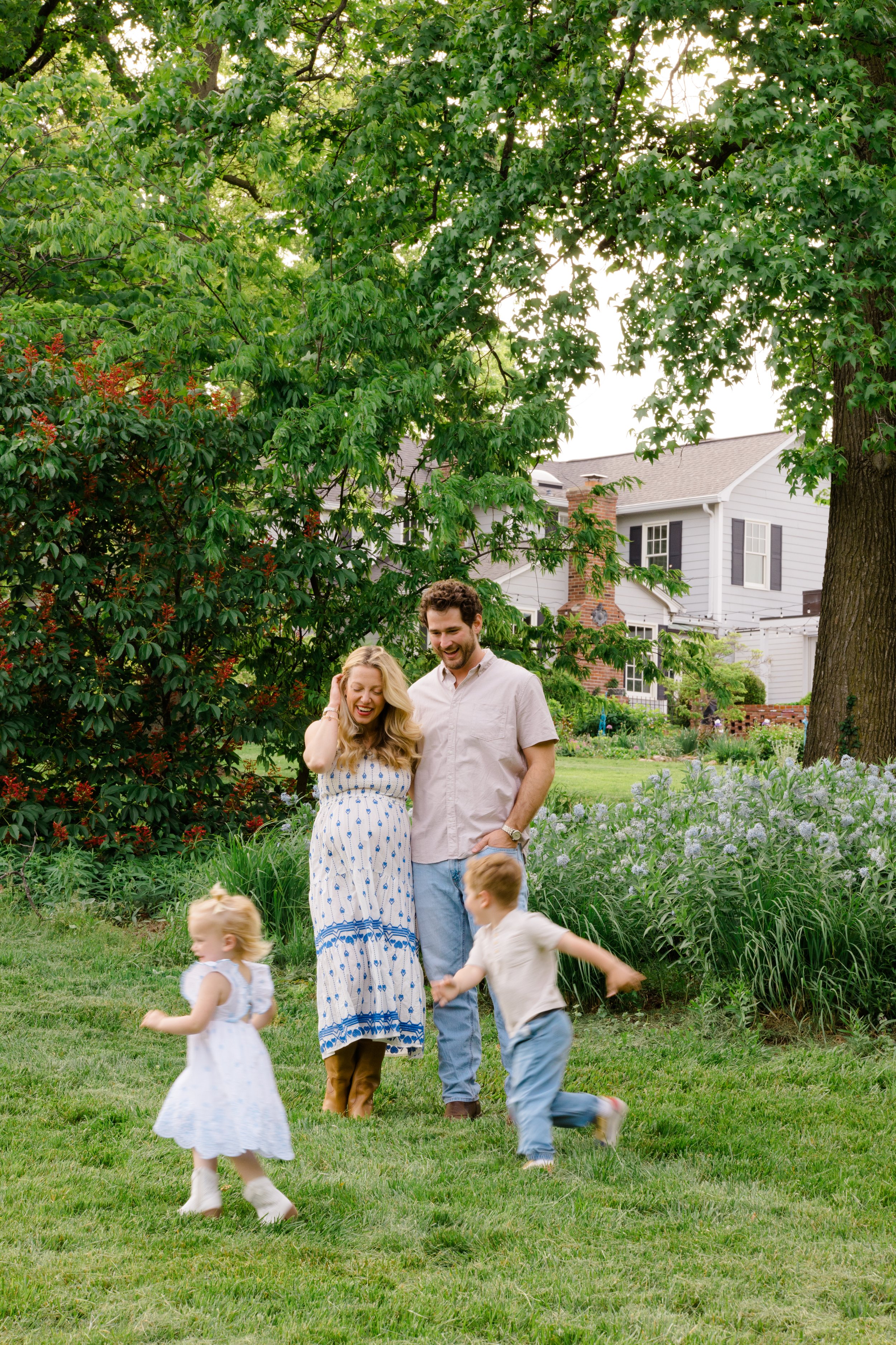 A family of four enjoying time in a backyard garden, with a woman, young girl, and two young boys playing on the grass amidst lush greenery and a white house in the background.