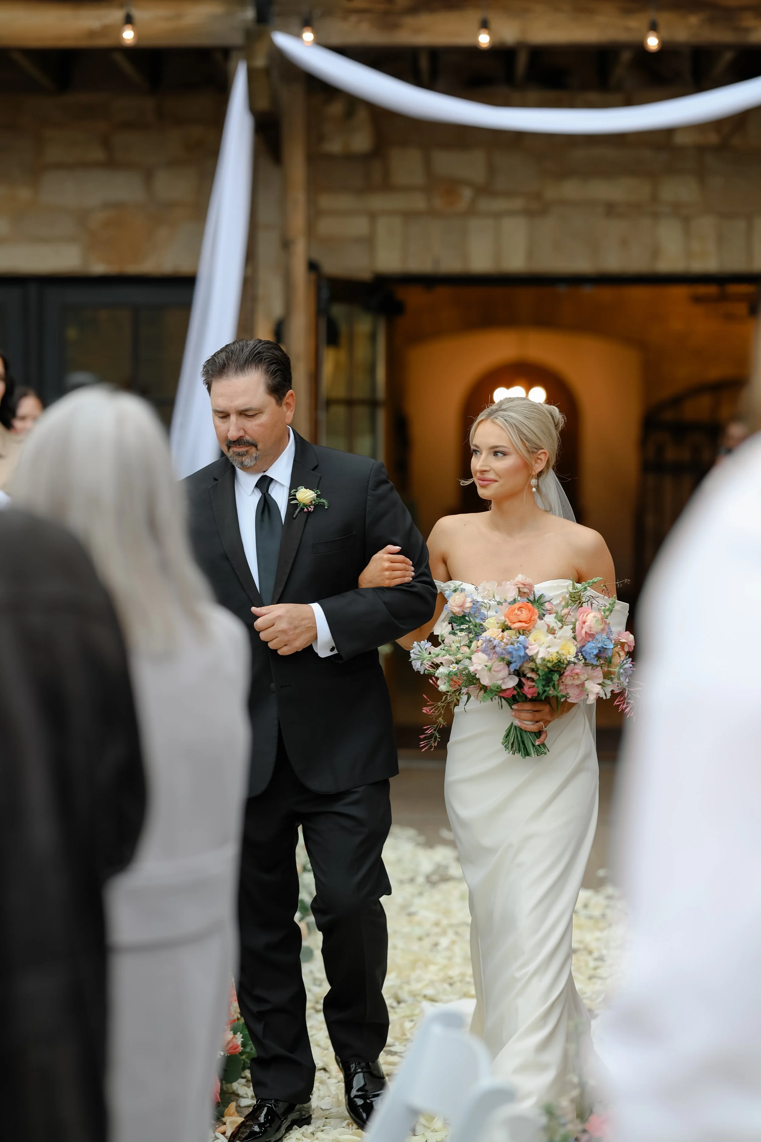 A bride in a strapless white wedding dress holding a colorful bouquet walking down an aisle with a man in a black suit and tie, inside a rustic venue with wooden beams and stone walls, during a wedding ceremony.