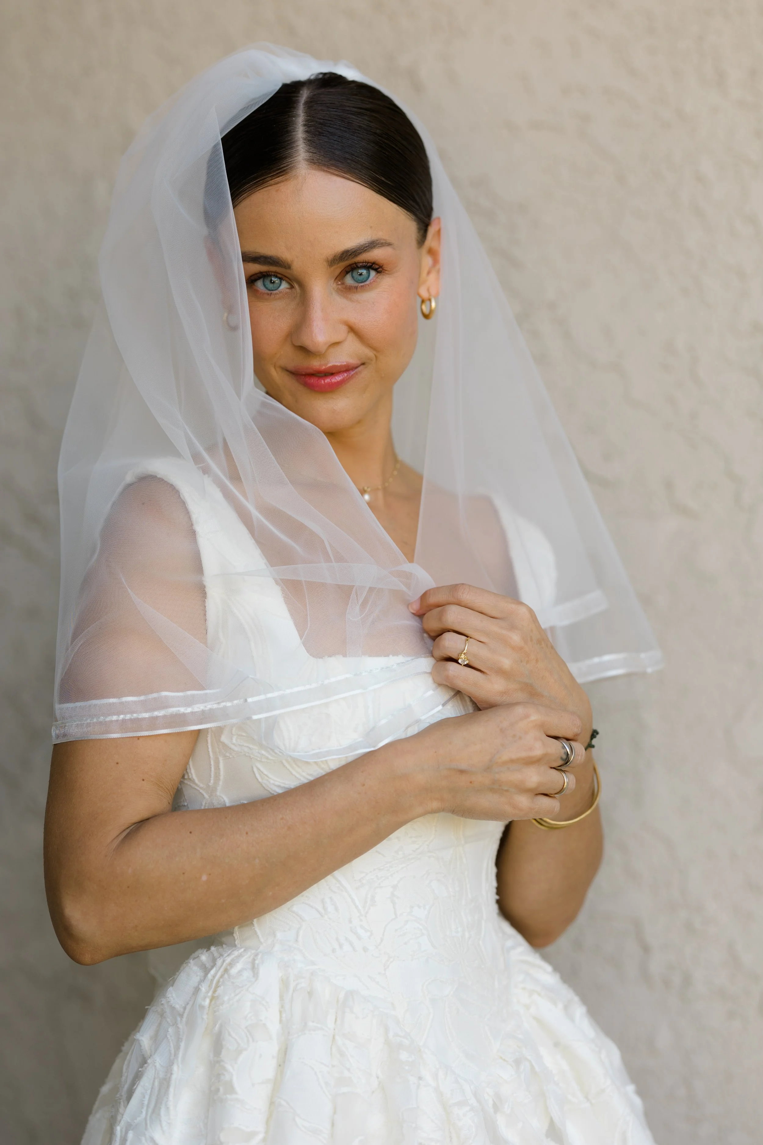 A woman in a white wedding dress with a sheer veil, holding the edge of her dress and smiling at the camera, standing against a plain beige wall.