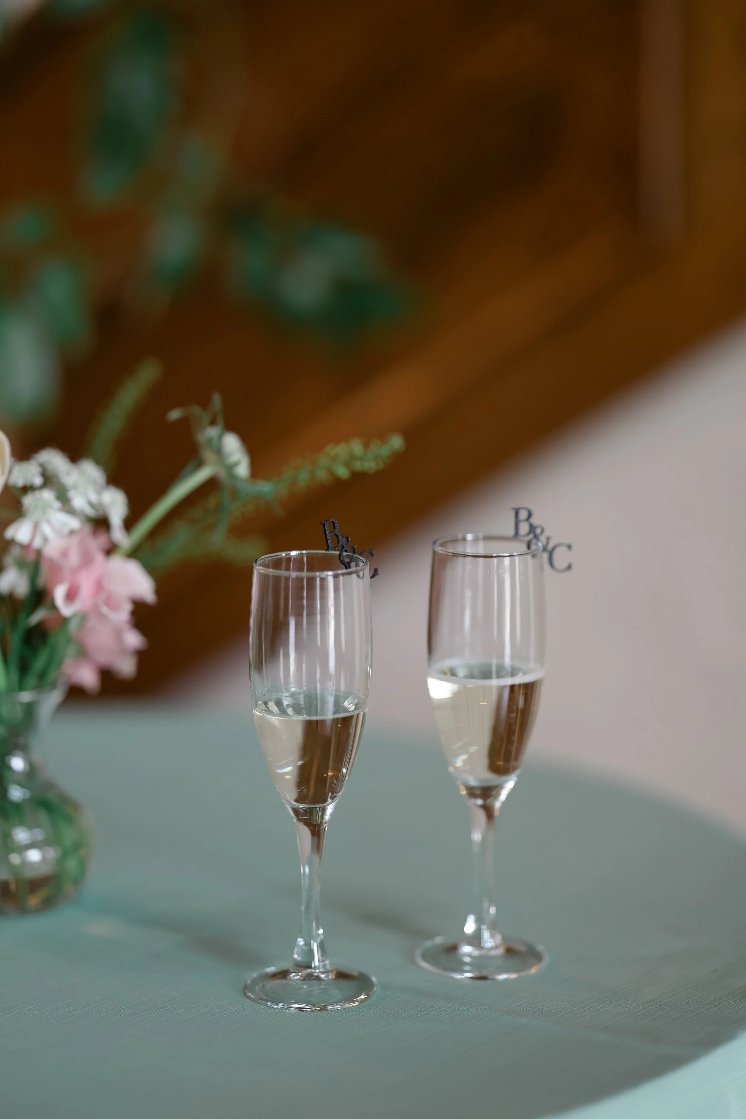 Three champagne glasses filled with bubbly champagne on a green table. Behind the glasses, there is a floral arrangement with pink and white flowers in a vase. In the background, a blurred wooden surface and some green foliage are visible.