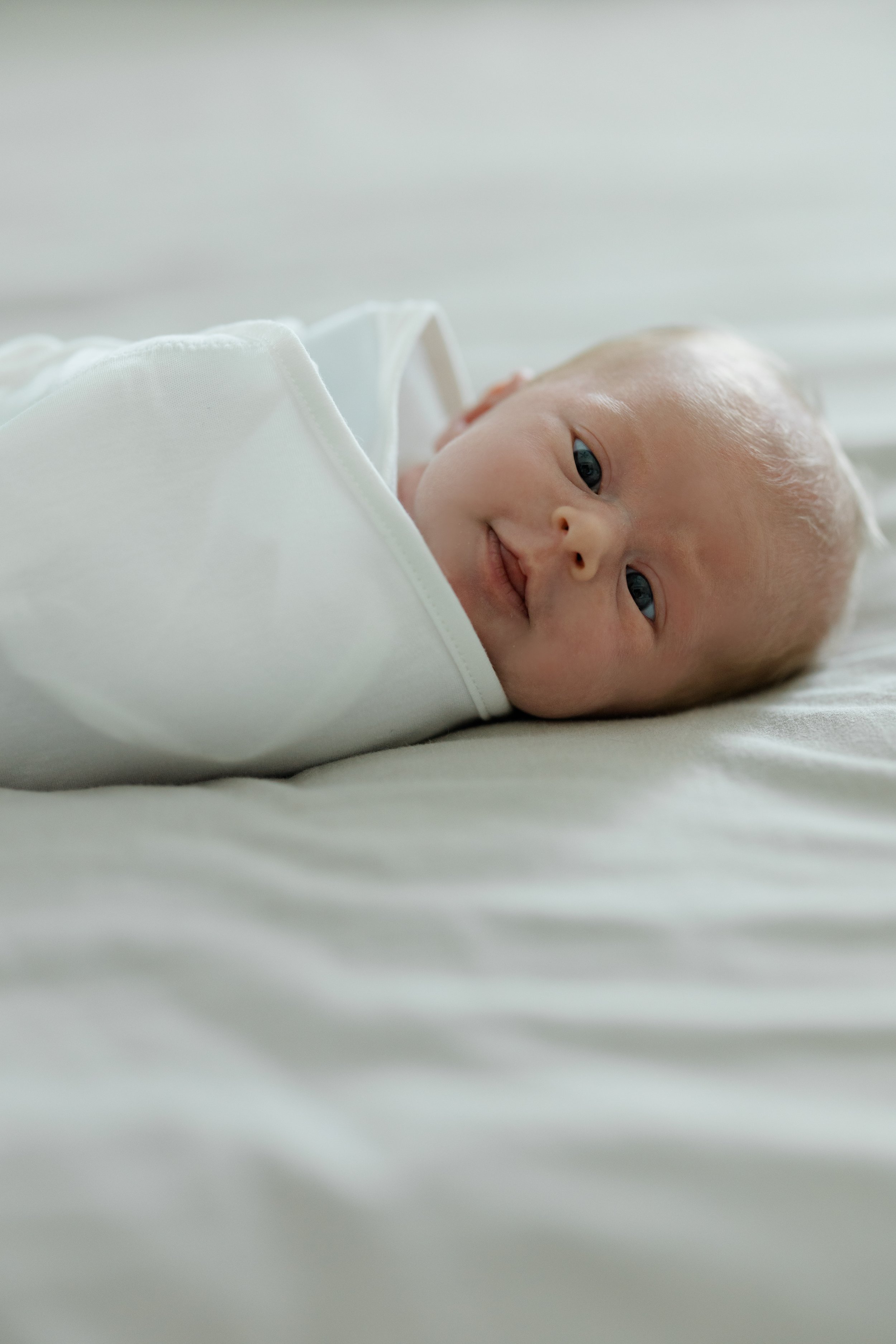 Close-up of a baby lying on a bed, wearing a white outfit, with blue eyes and light-colored hair, looking at the camera with a slight smile.