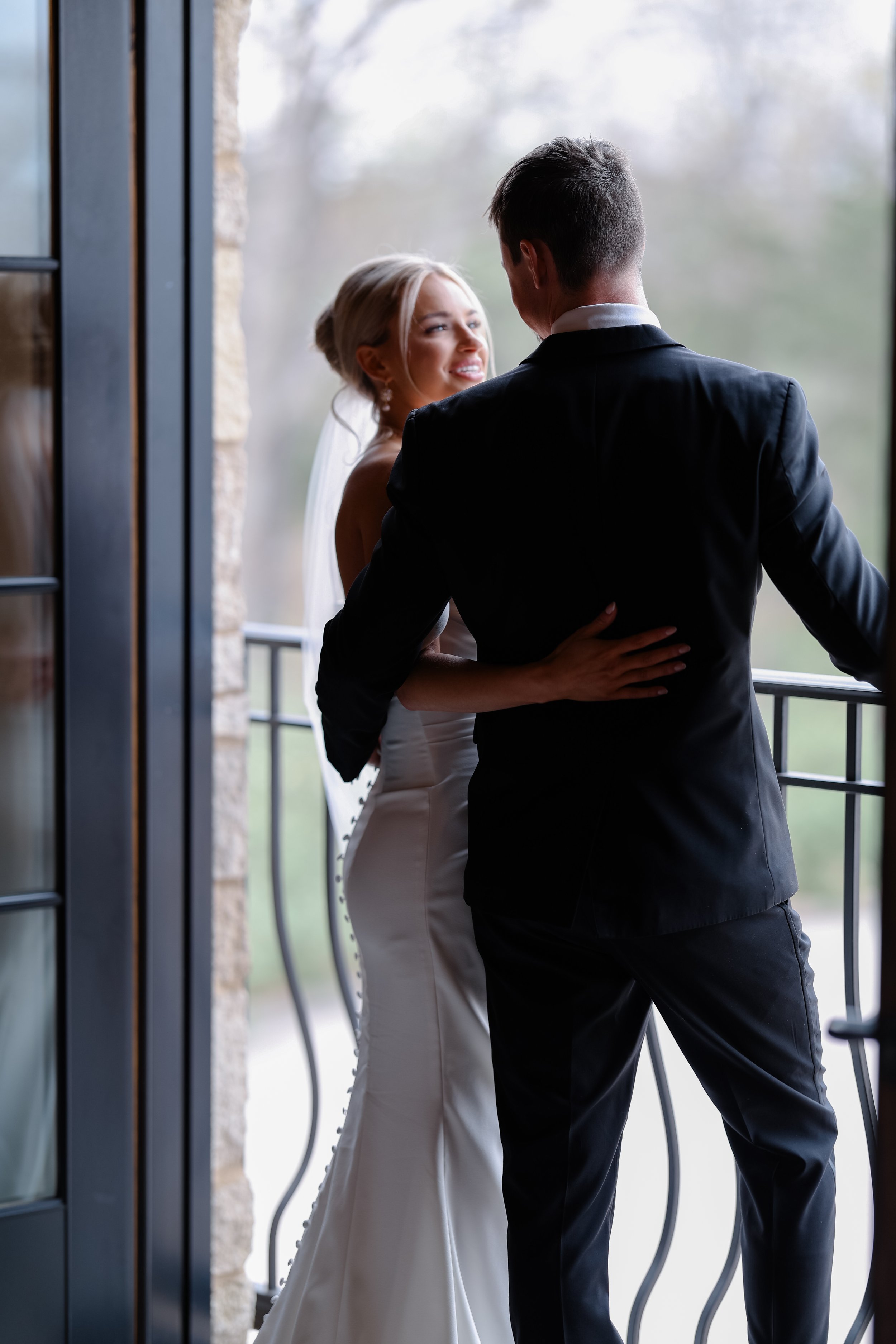 A bride and groom facing each other near a doorway, smiling, with outdoor scenery in the background.