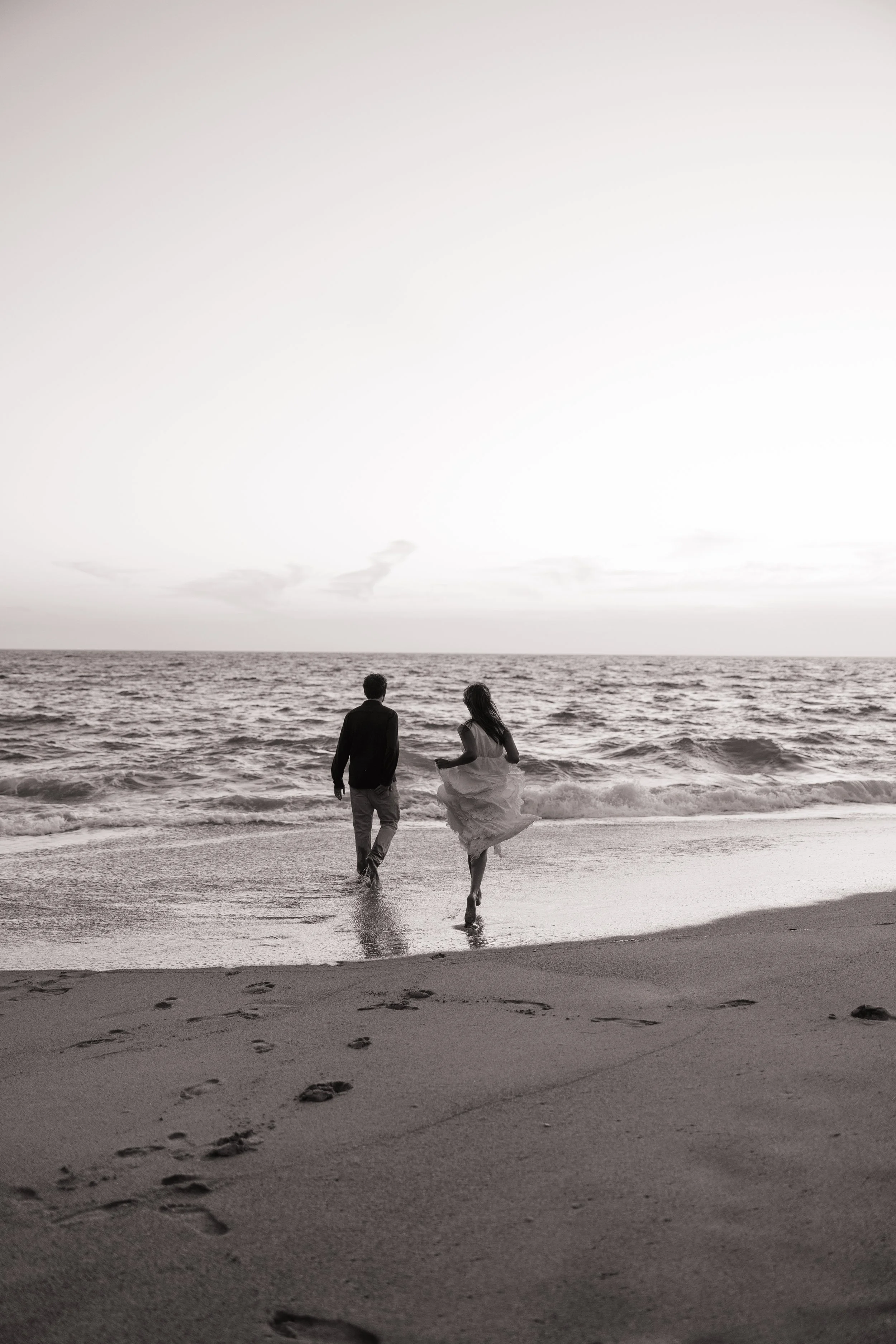 A black and white photo of a couple walking along the shoreline at sunset or sunrise, leaving footprints in the wet sand, with the ocean and sky in the background.