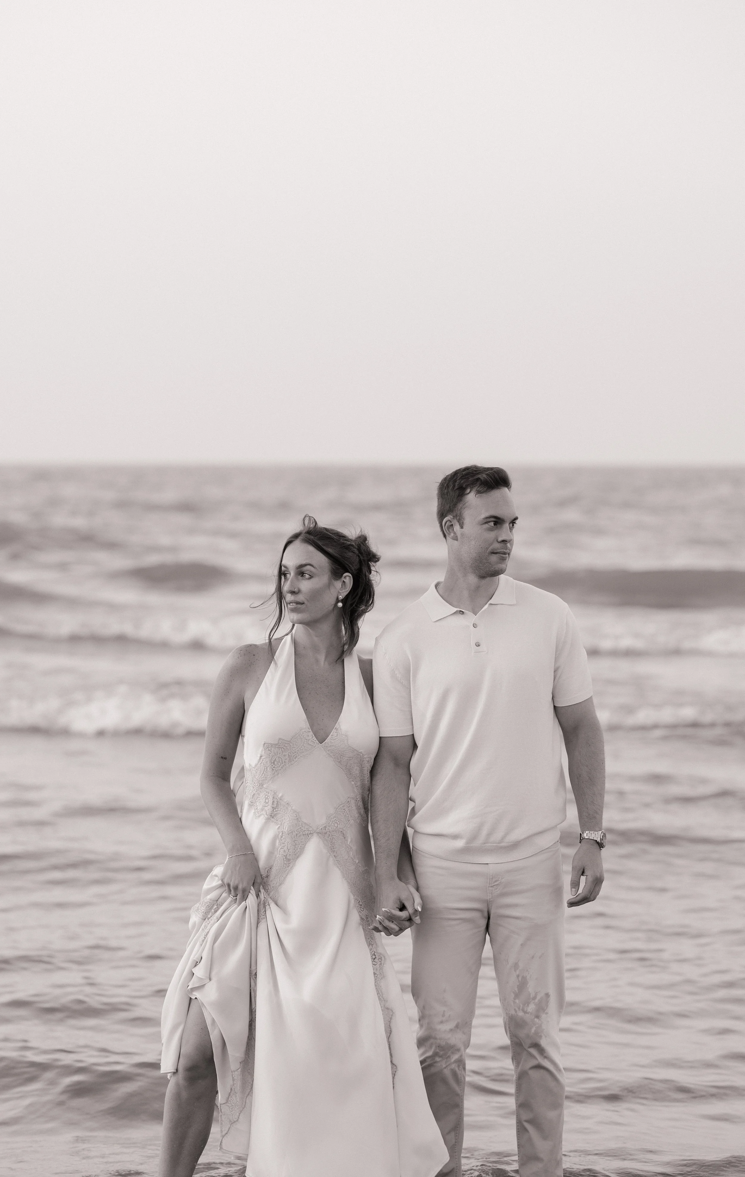 A black-and-white photo of a couple walking along the beach holding hands, with the ocean in the background.