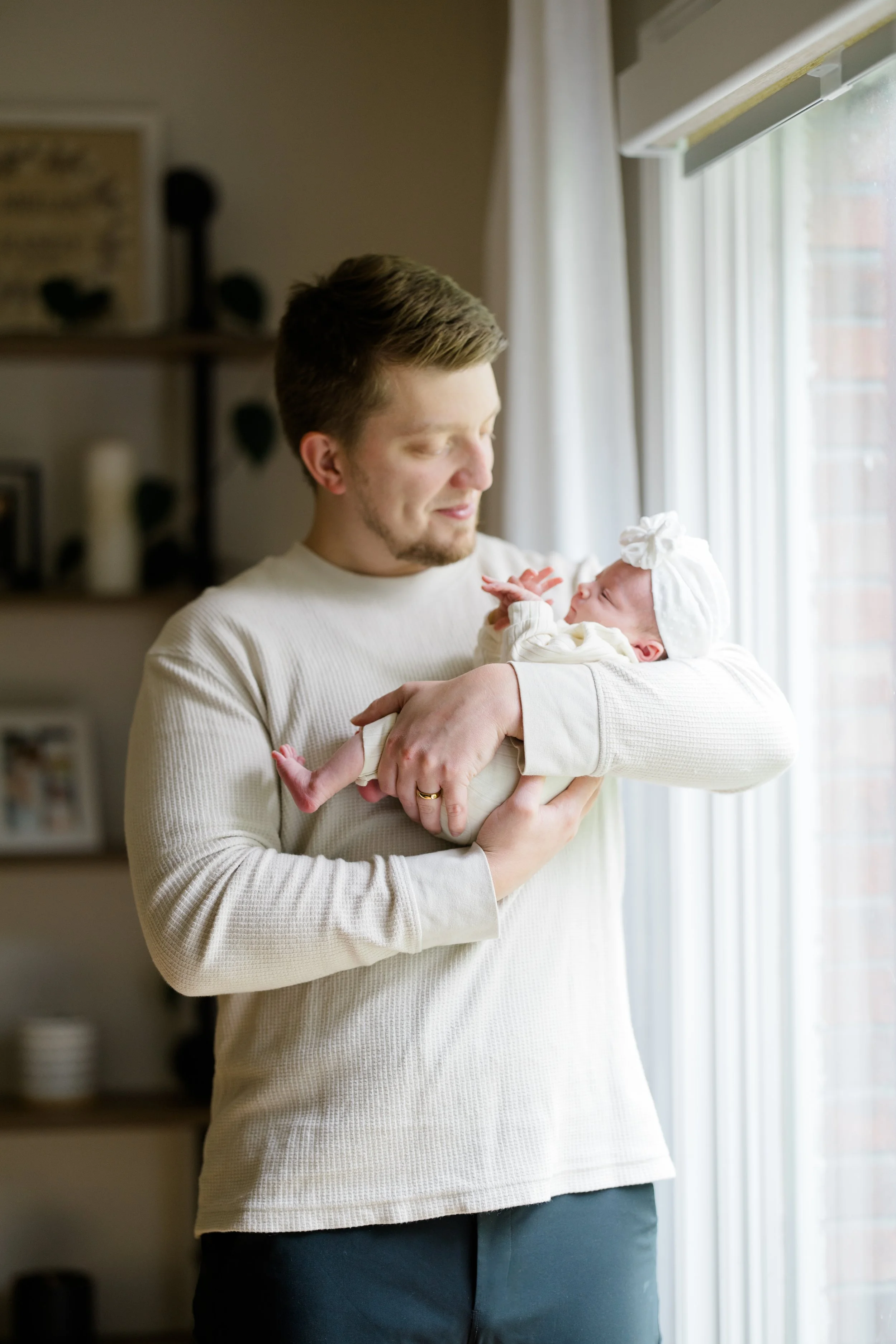 A man holding a newborn baby near a window with white curtains, smiling and gazing at the baby.