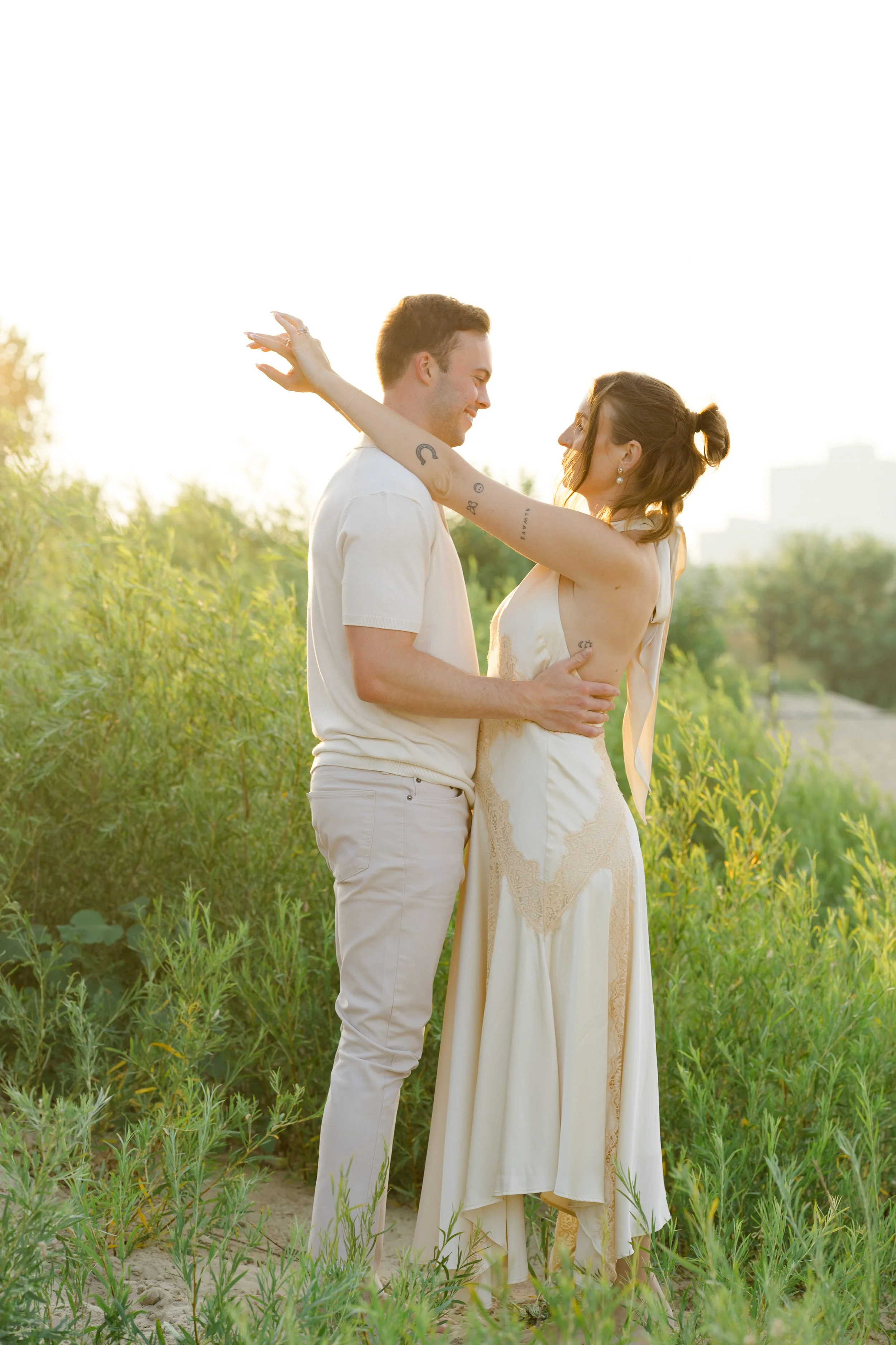 A man and woman standing closely in a field of green plants, smiling at each other during sunset, with the woman extending her arms around the man's neck.
