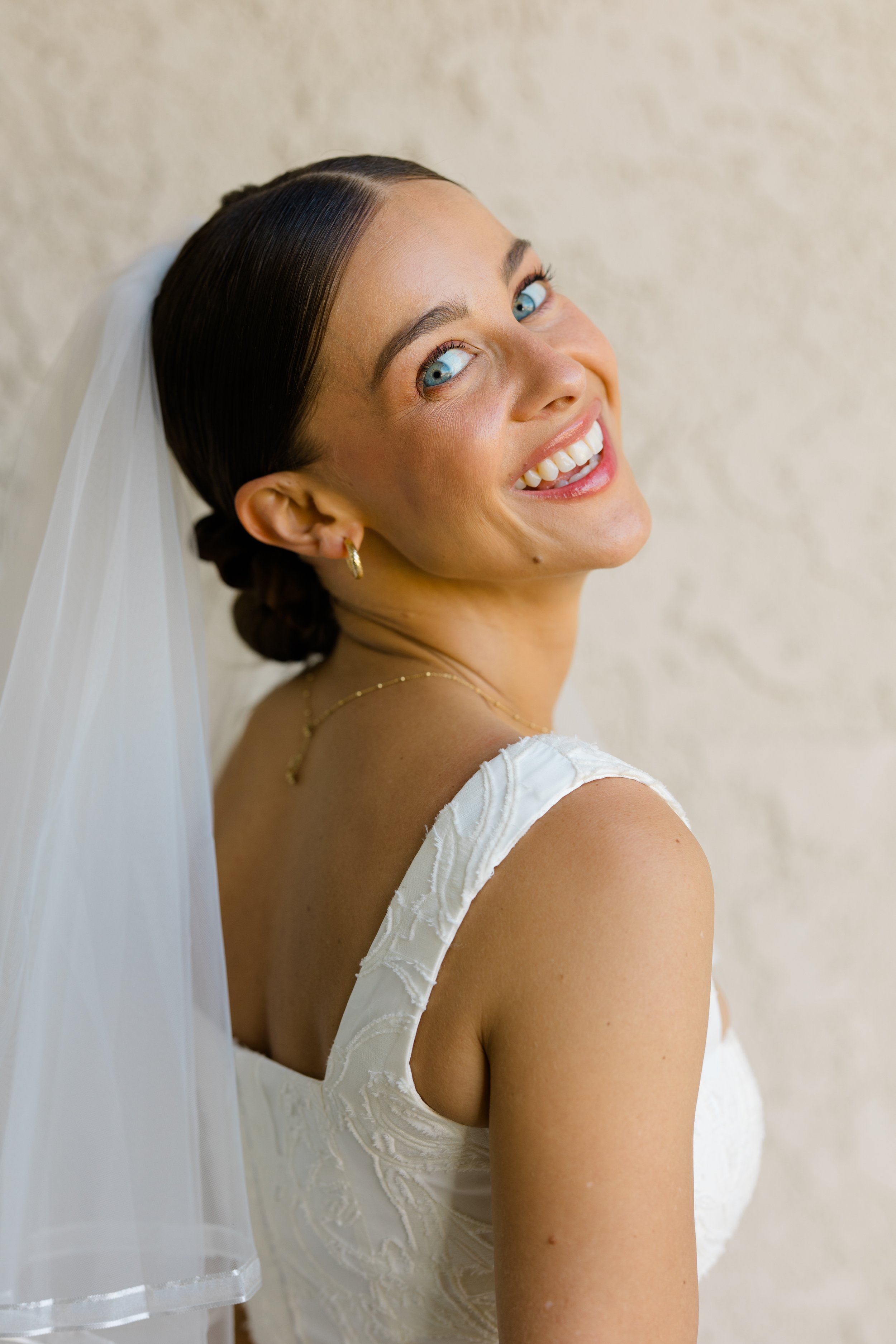 Close-up of a smiling woman with blue eyes, dark hair styled back, wearing a white sleeveless dress with embroidery, gold hoop earrings, a delicate gold necklace, and a sheer white veil, against a beige background.