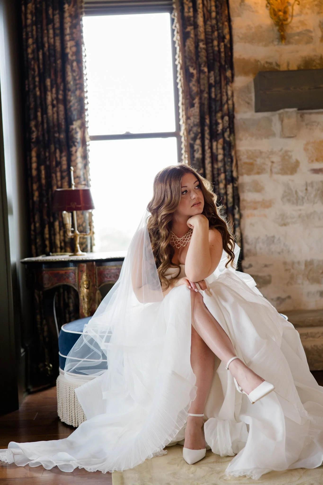 A bride in a white wedding gown sitting on a stool, with her hand resting on her chin, in a room with stone walls and large windows with patterned curtains.
