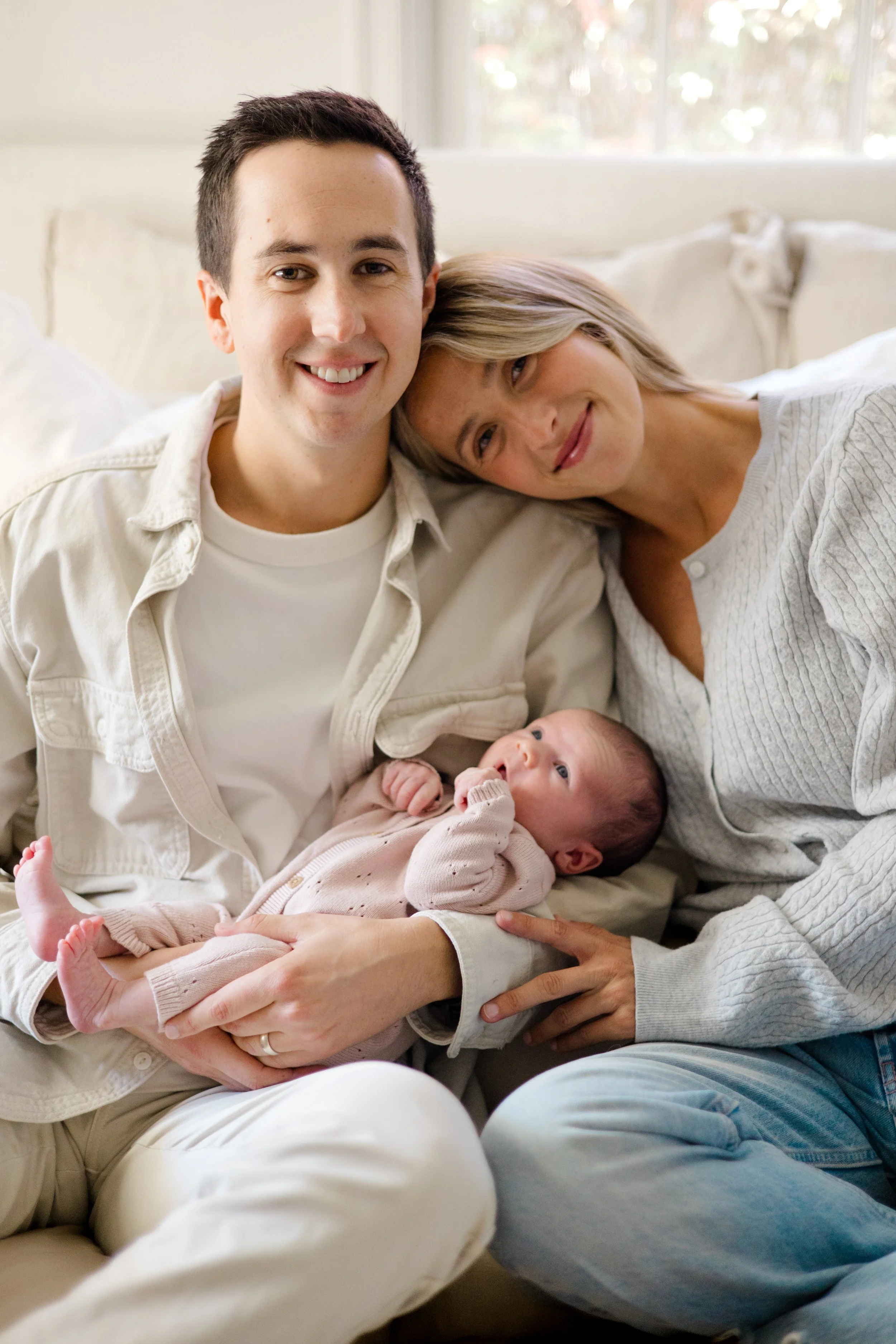A smiling young man, a woman leaning her head on his shoulder, and a baby lying on their lap, all sitting on a sofa in a bright room.