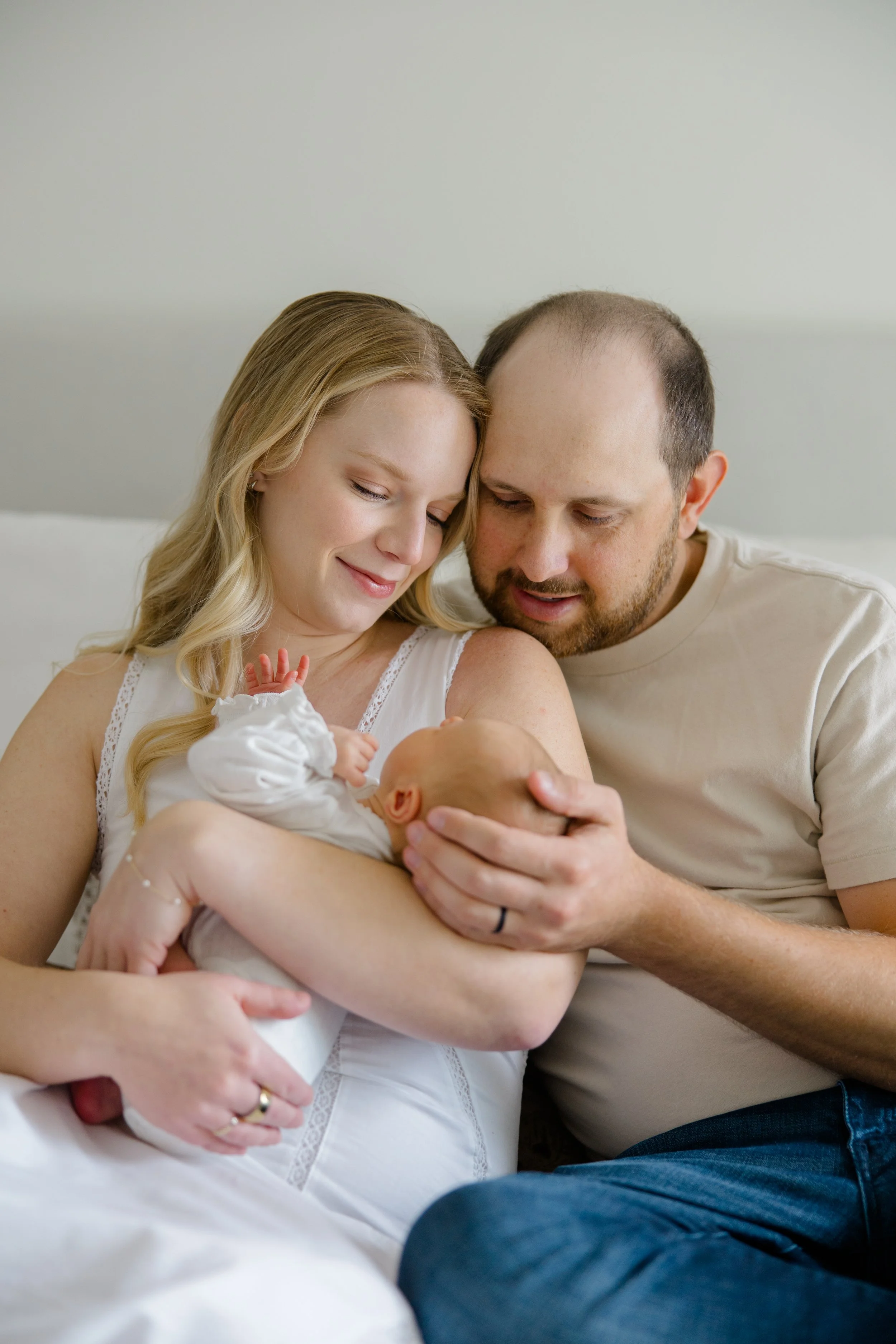 A woman and a man holding a newborn baby, smiling and looking at the baby, in a room with a plain background.