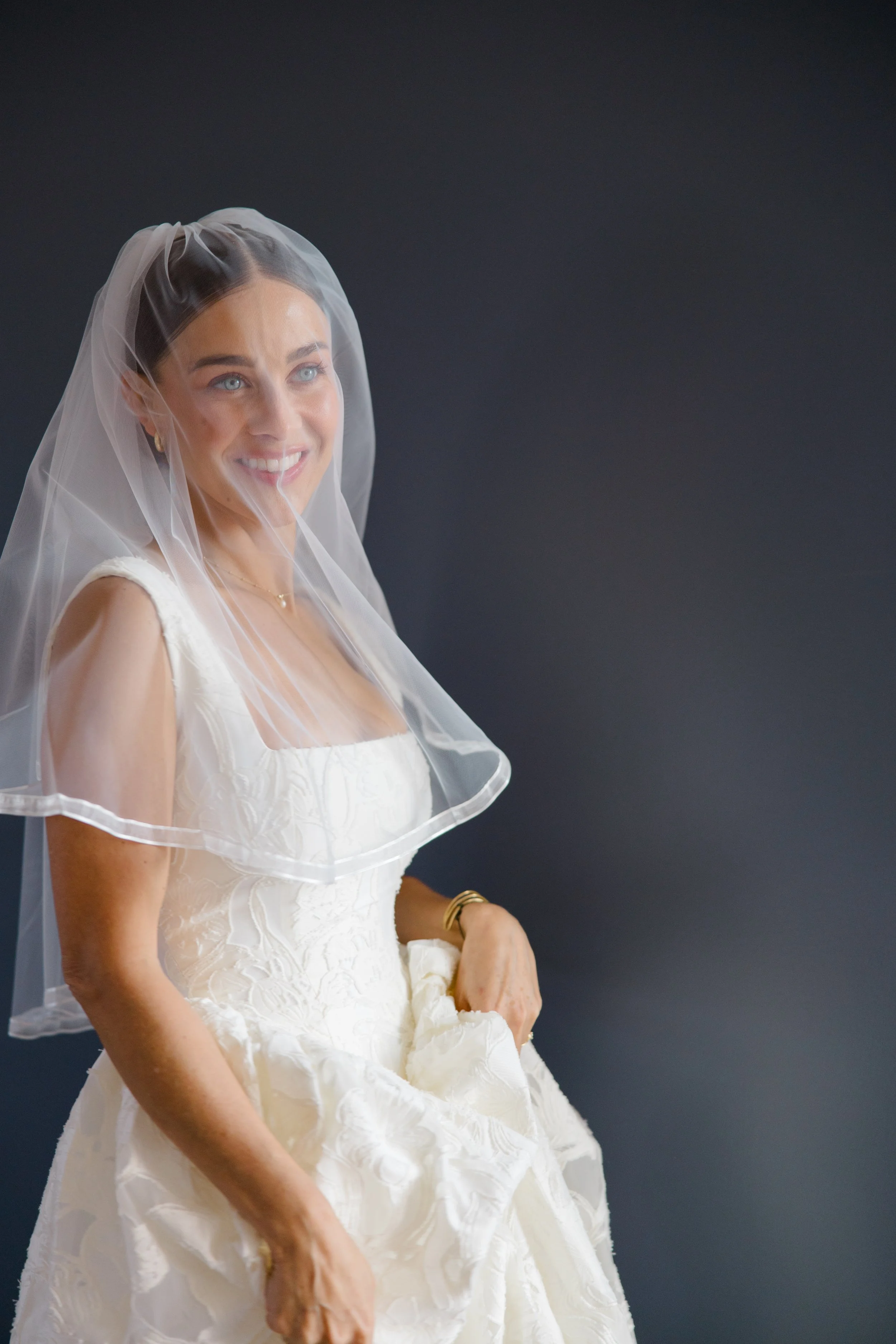 A smiling woman in a white wedding dress with a sheer veil on her head, standing against a dark background.