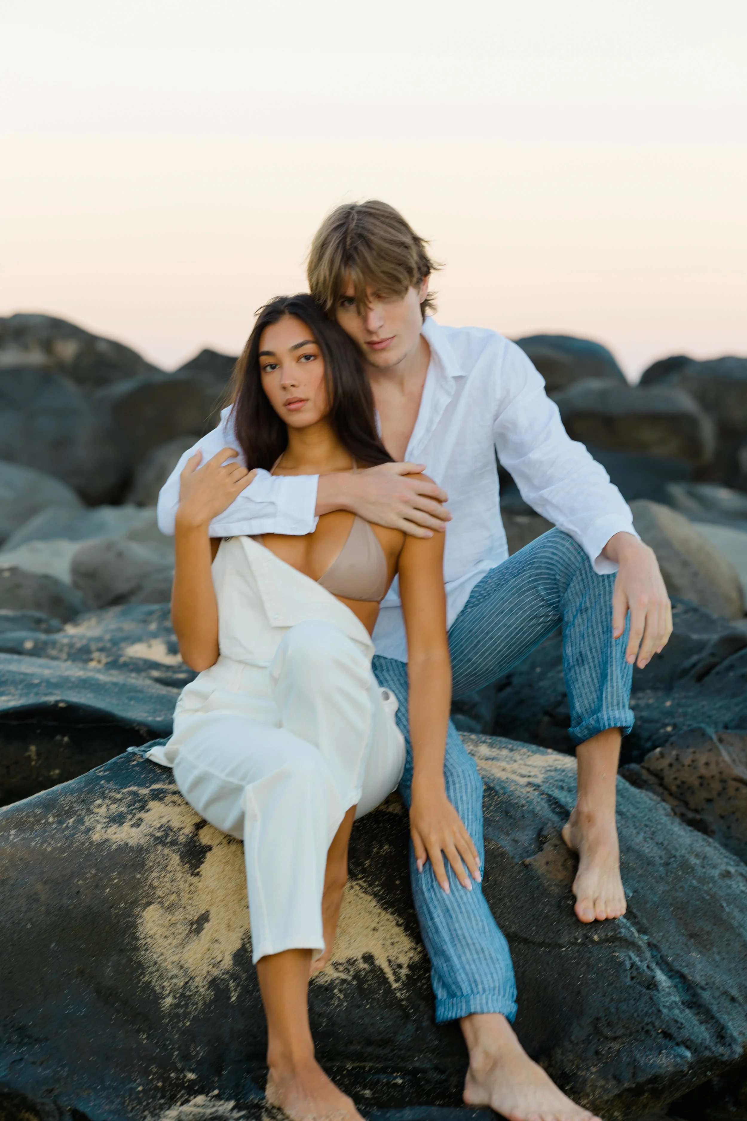 A young couple sitting close together on rocks by the water at sunset, looking into the camera. The woman has long dark hair and is wearing a beige top and white pants. The man has light brown hair and is wearing a white shirt and blue striped pants.