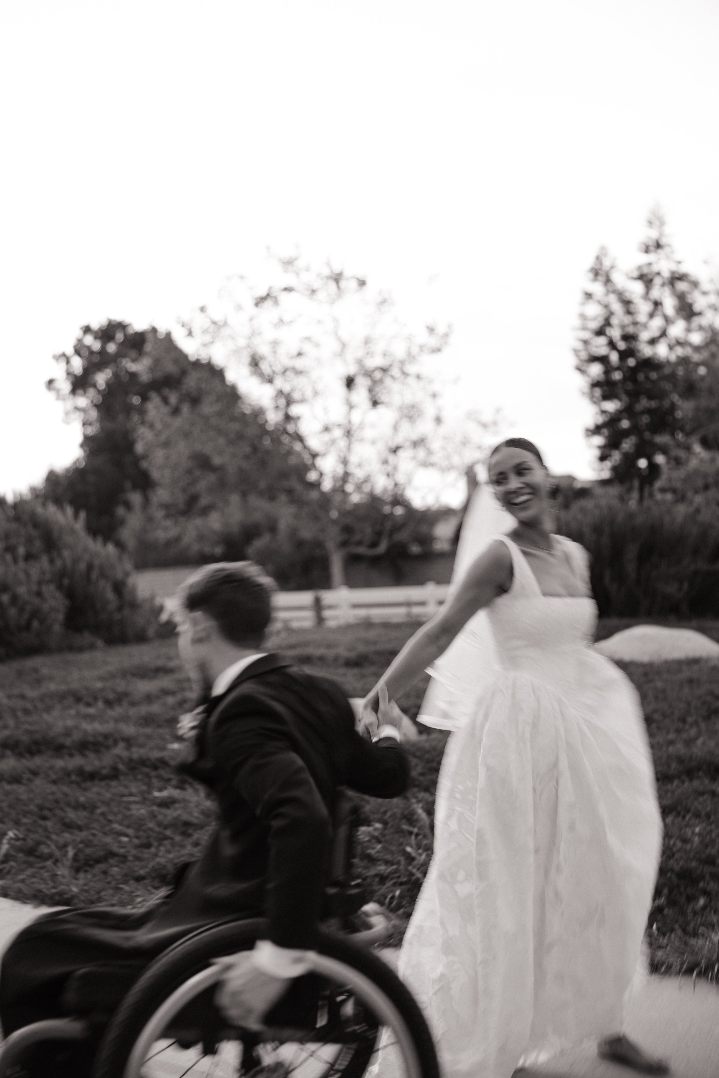 A woman in a white wedding dress holding hands with a man in a wheelchair outdoors in a park with trees in the background. The woman is smiling, and the scene appears joyful.