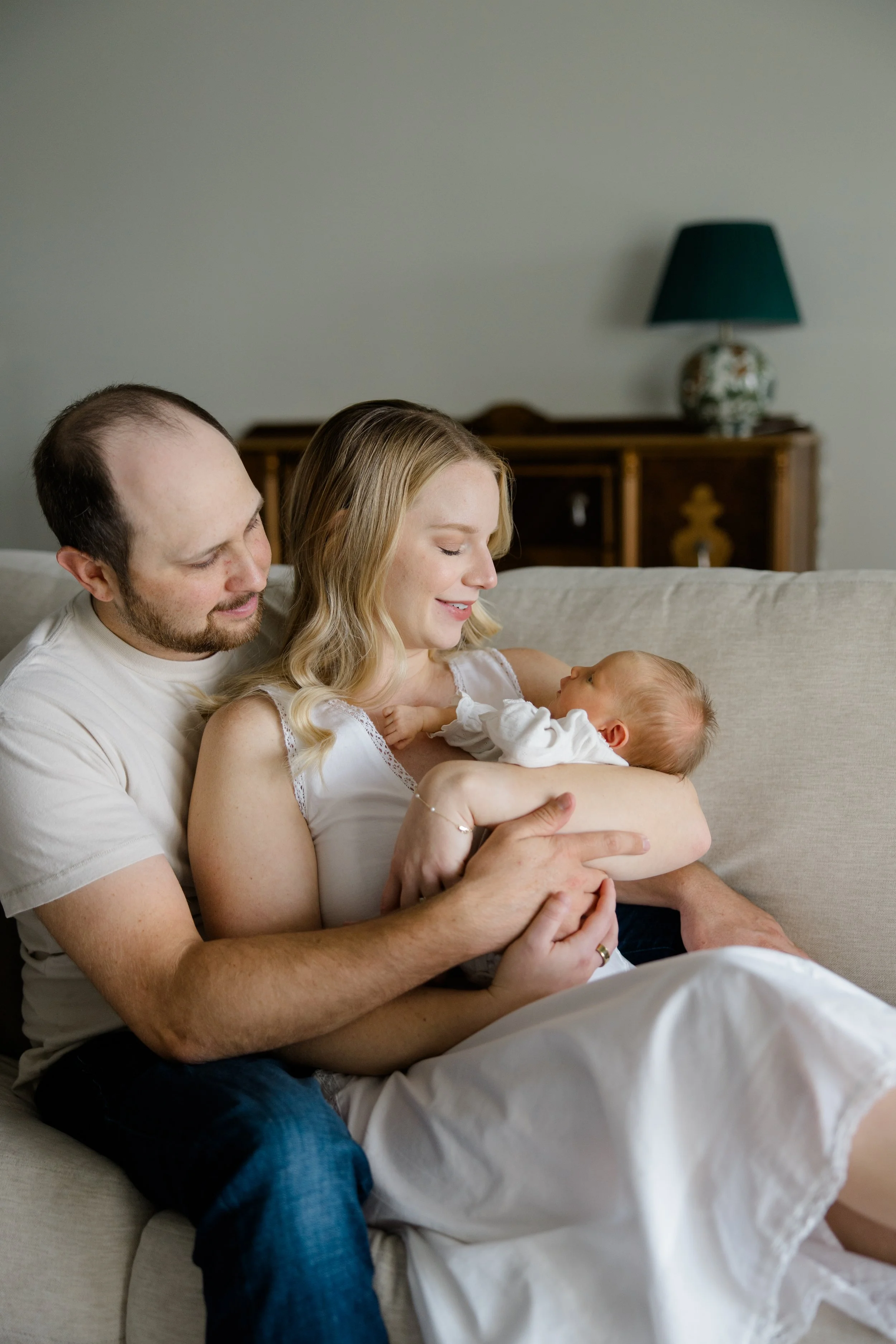 A family sitting on a couch with a woman holding a baby, a man sitting close and smiling, in a room with a wooden sideboard and a table lamp in the background.
