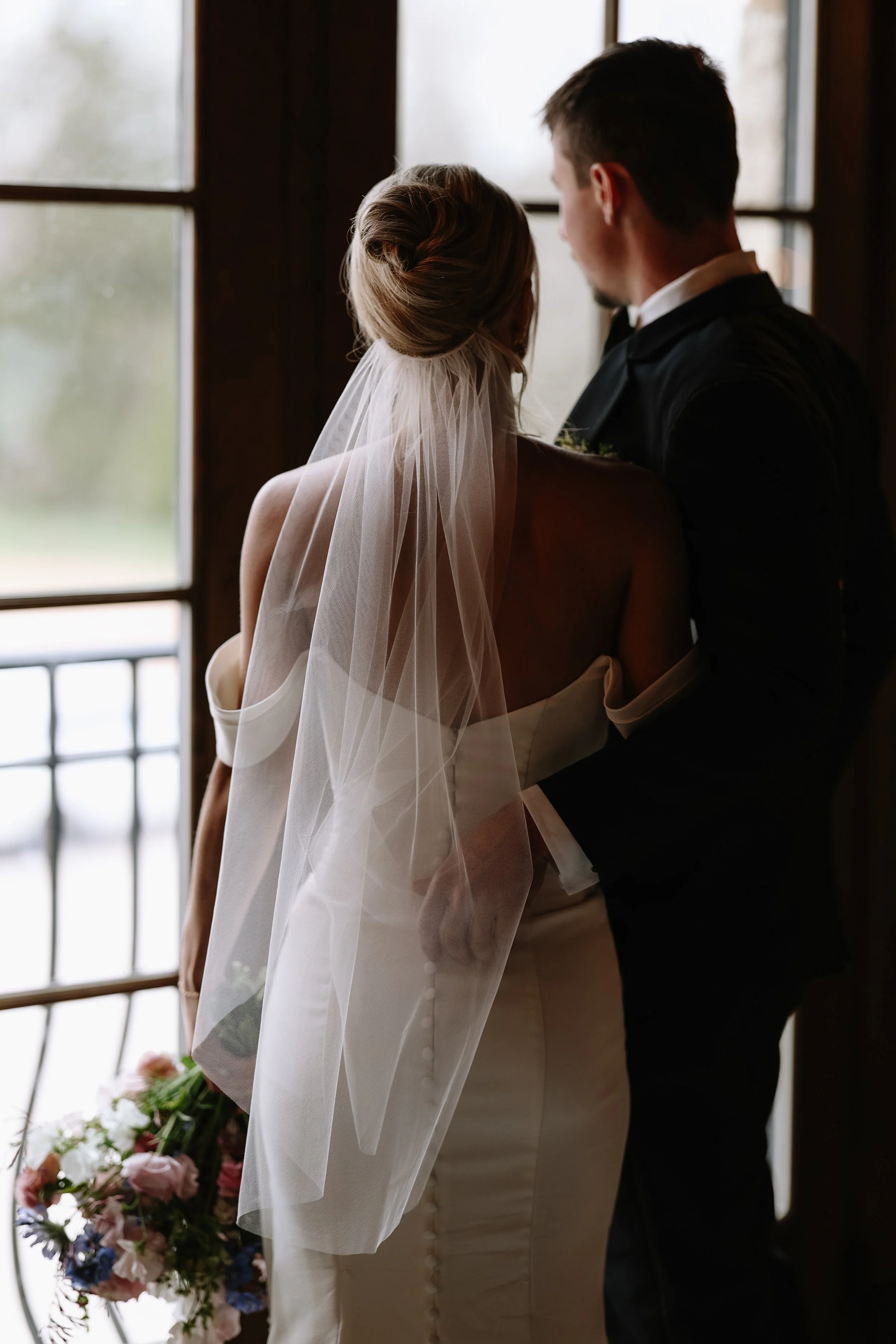 A bride with a veil and a groom in a tuxedo share a moment by a window, with a bouquet of flowers in the background.