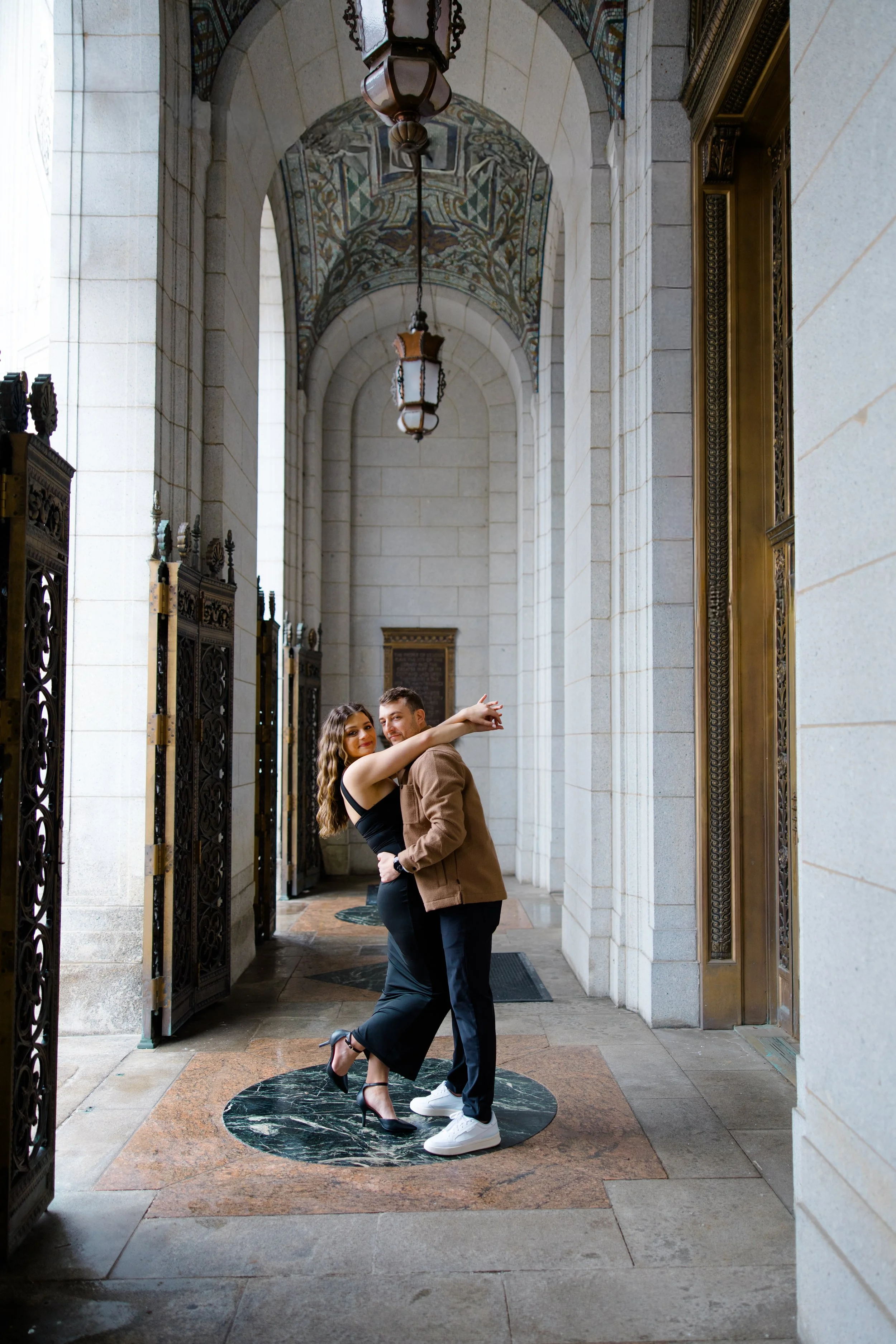 A man and woman dancing together in a covered outdoor corridor with ornate lanterns, arched ceilings with colorful mosaic patterns, and decorative metal gates.
