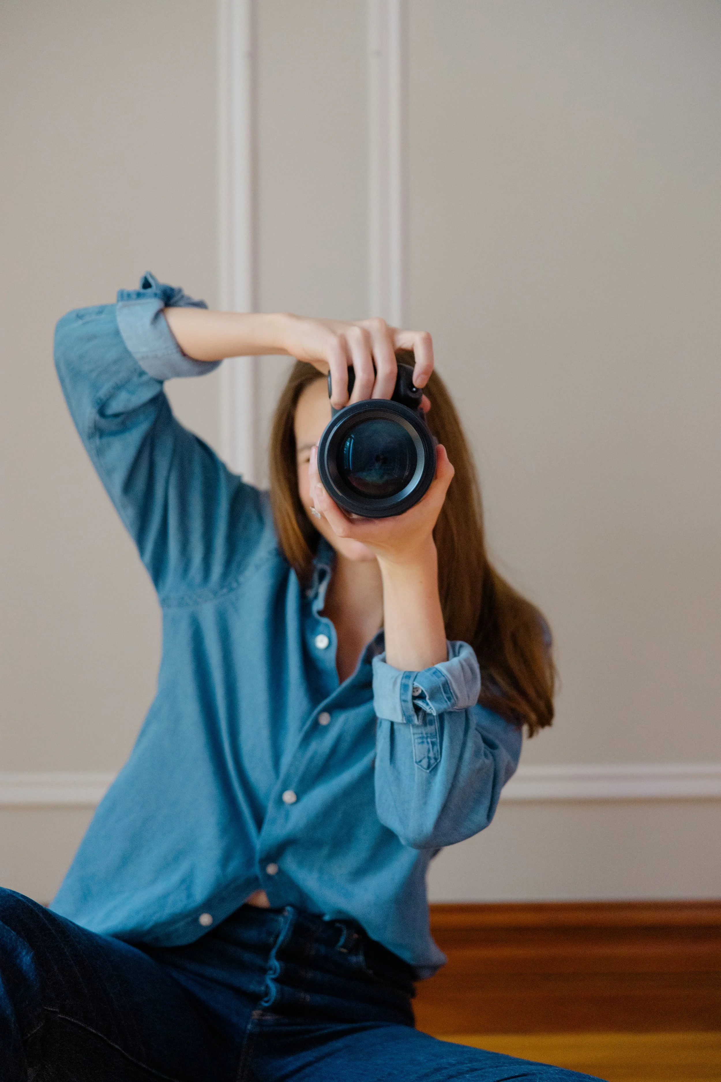 Person sitting on the floor taking a photo with a camera, wearing a blue denim shirt and jeans, with a plain wall background.