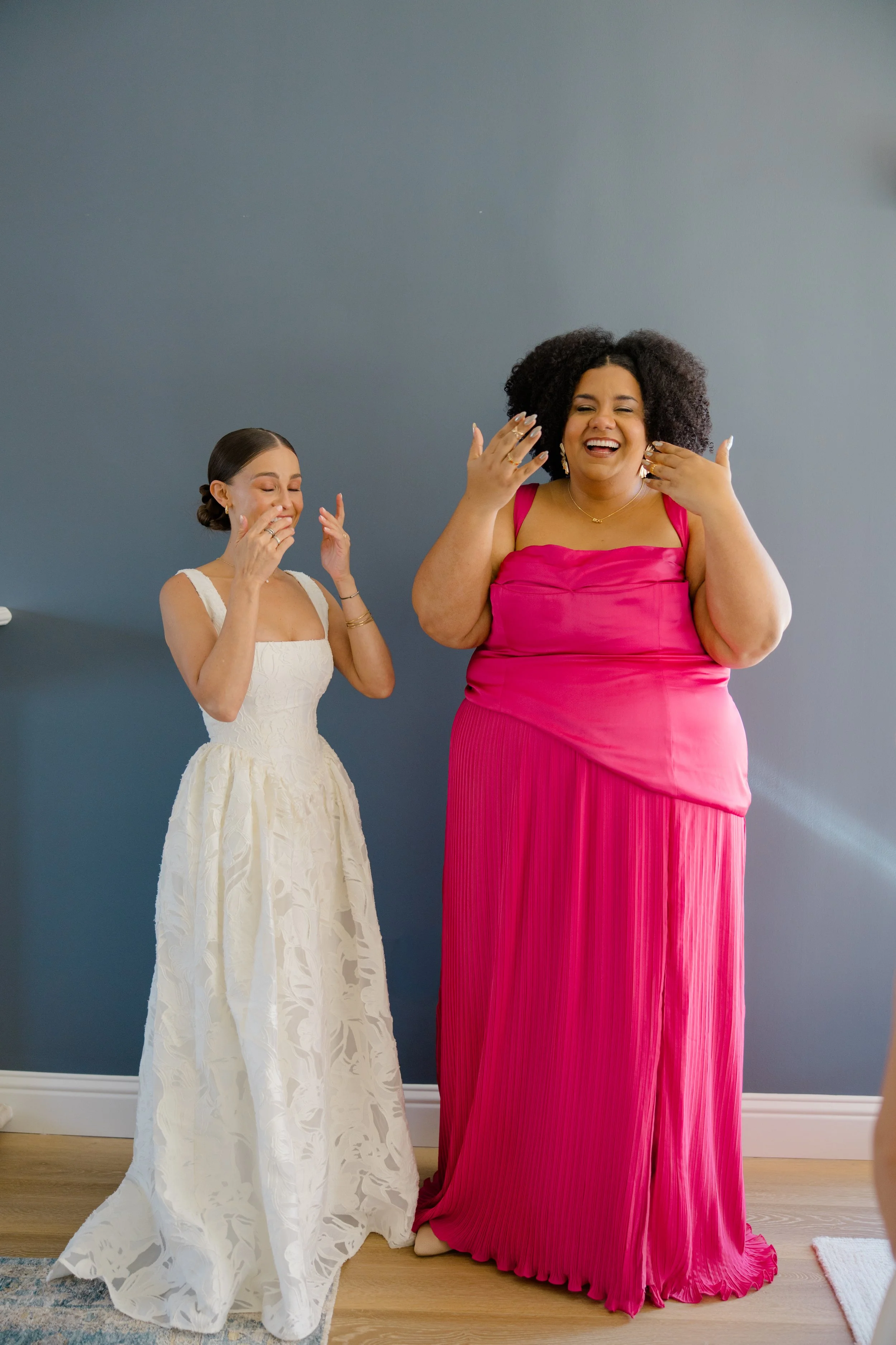 Two women are laughing joyfully, one in a white lace dress and the other in a bright pink dress, standing in a room with a blue wall and wooden floor.