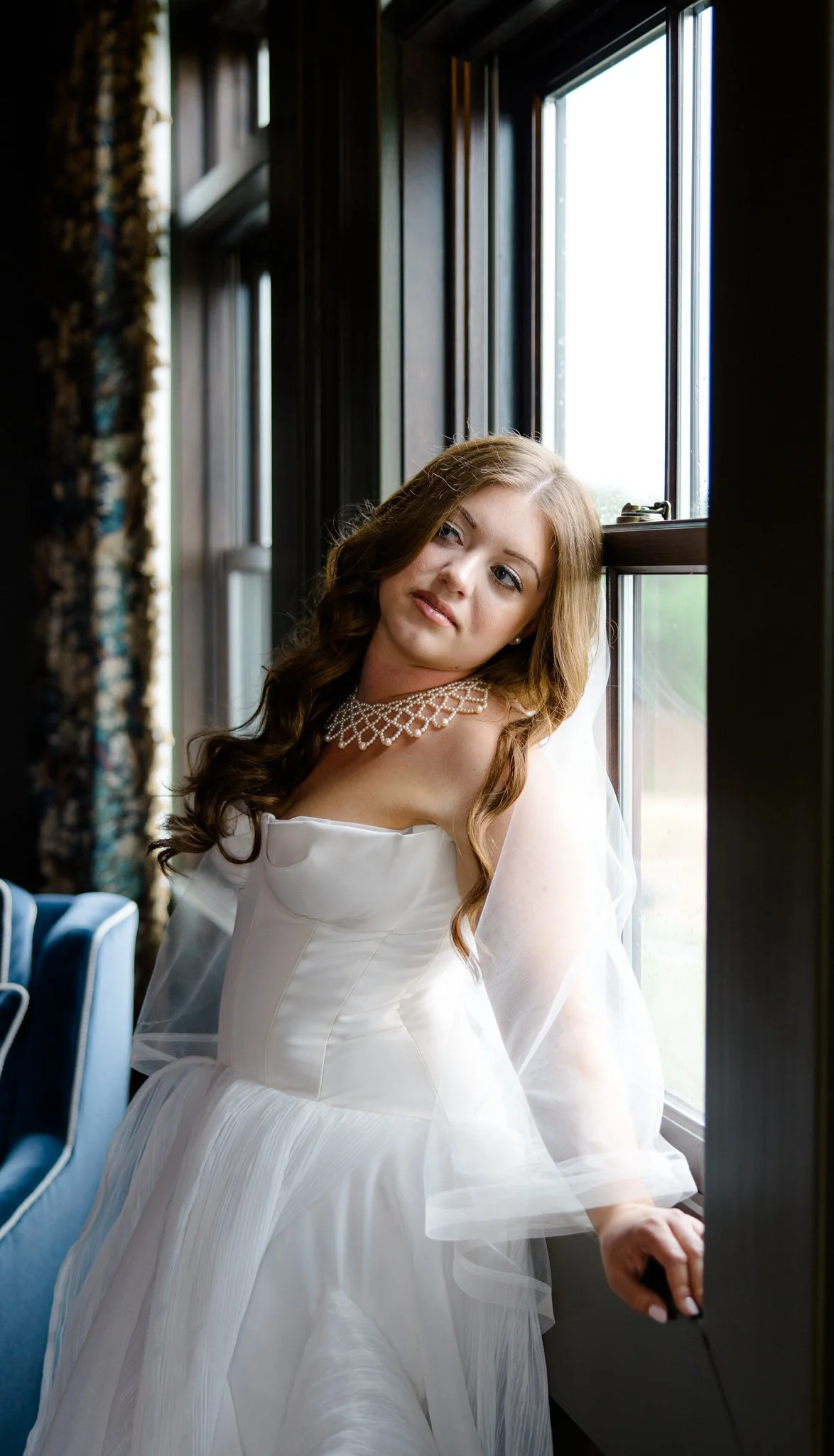 A young woman with long, wavy brown hair wearing a white dress and a pearl necklace, sitting by a window with natural light coming in, inside a room with dark wood paneling.