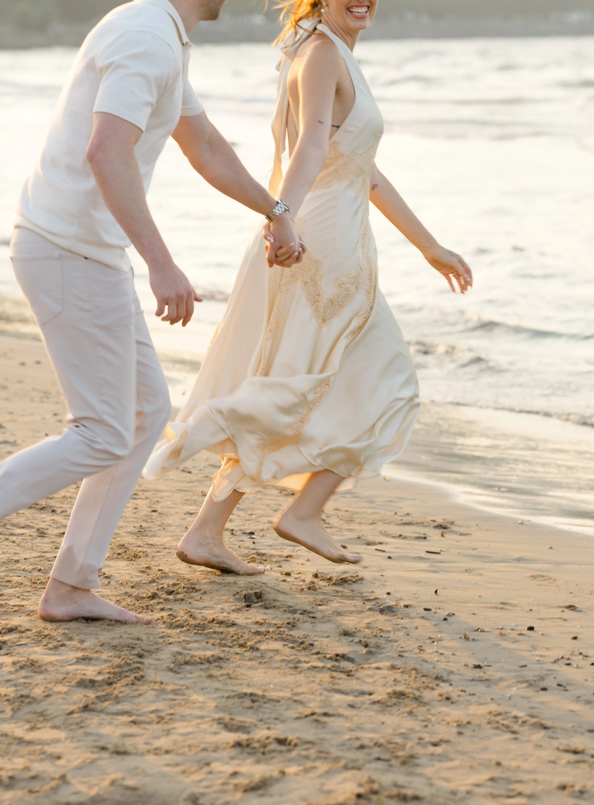 A couple holding hands and walking barefoot on the beach at sunset, with the woman smiling.