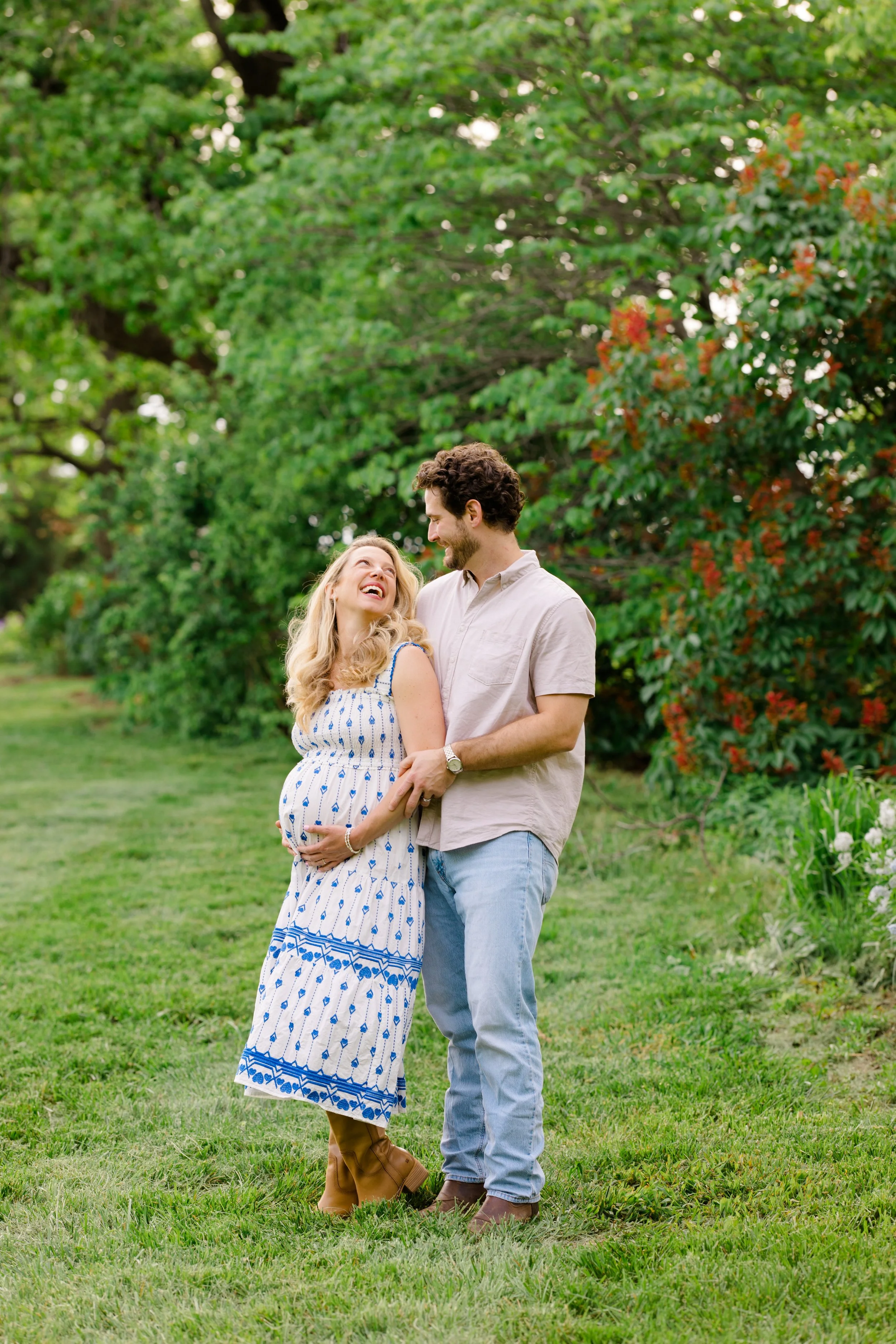 A pregnant woman and a man standing together outdoors, smiling and looking at each other, surrounded by green trees and grass.