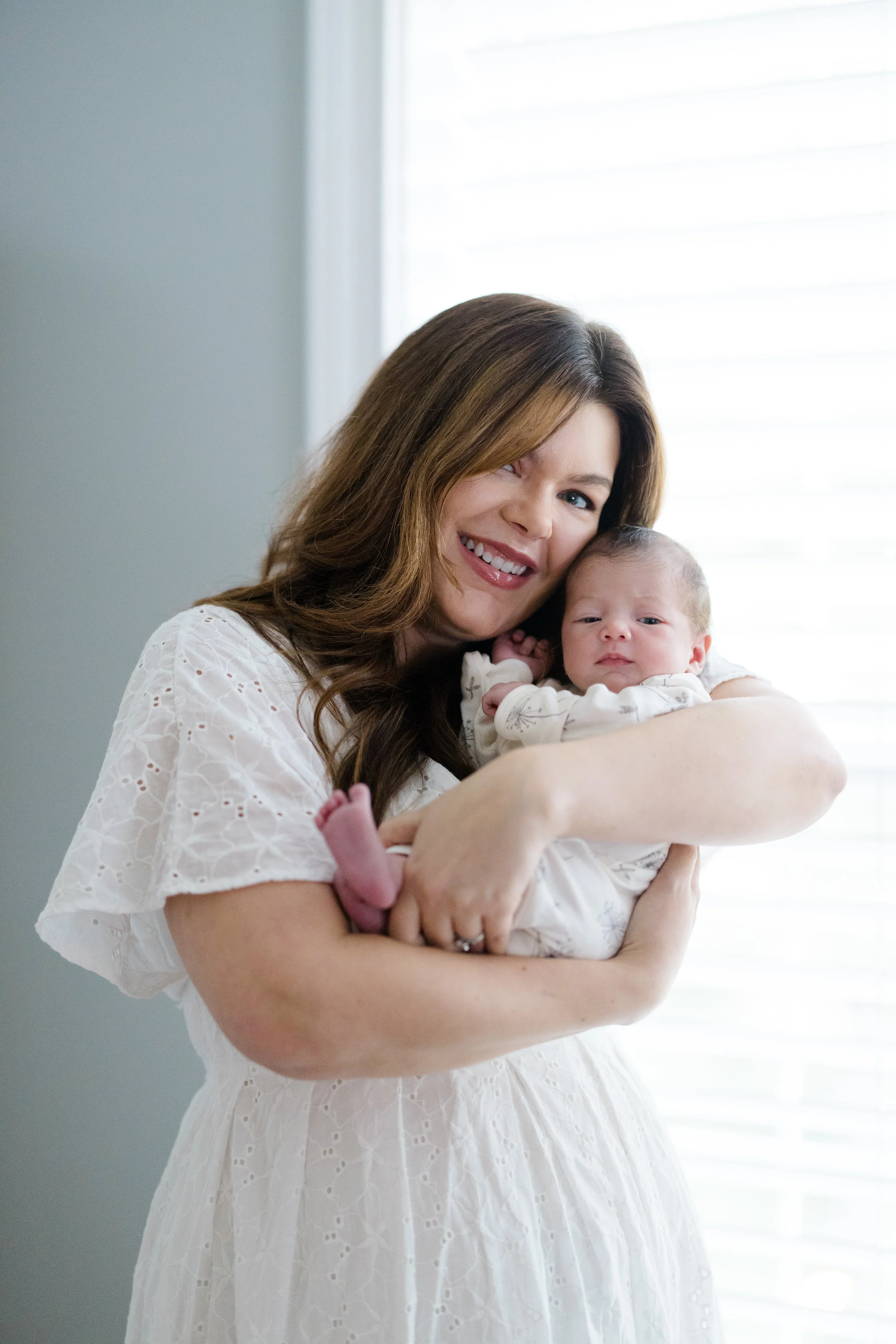 A woman with long brown hair, wearing a white dress, smiling, holding a newborn baby in a white outfit, standing near a window with white blinds.