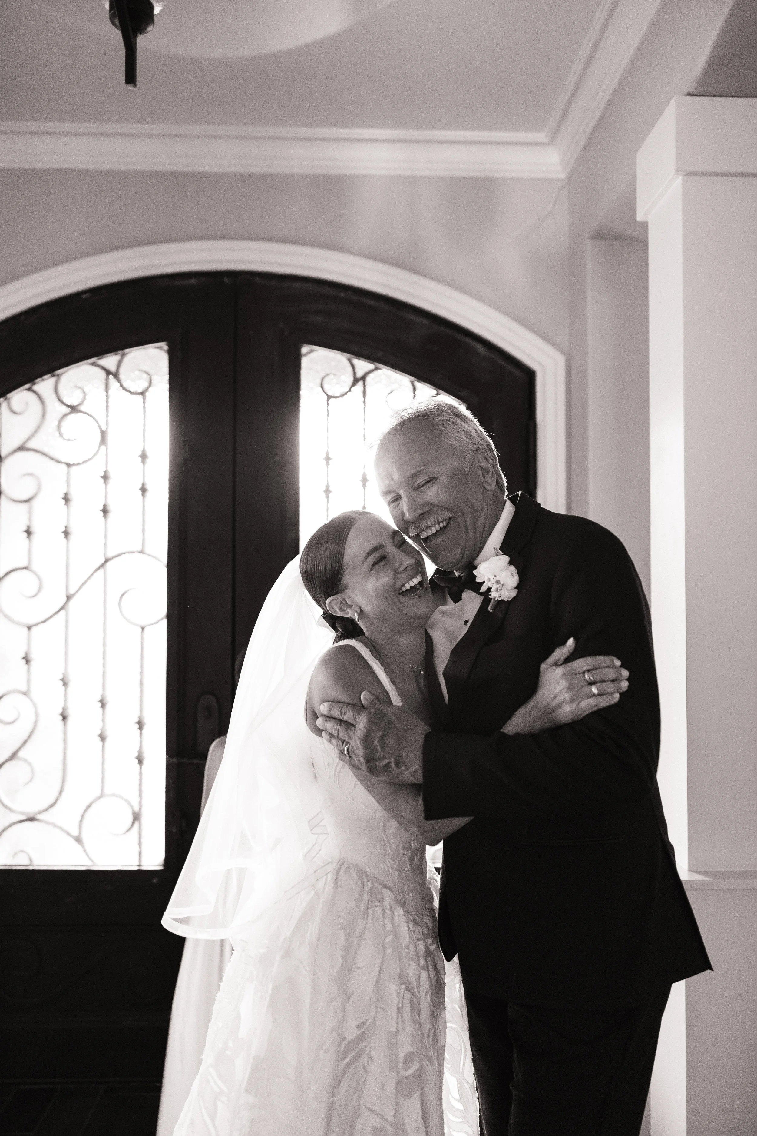 An elderly man and a woman in a wedding gown are embracing and smiling happily inside a house near a door with iron details. The man is wearing a tuxedo with a boutonniere, and the woman is wearing a lace wedding gown with a veil.