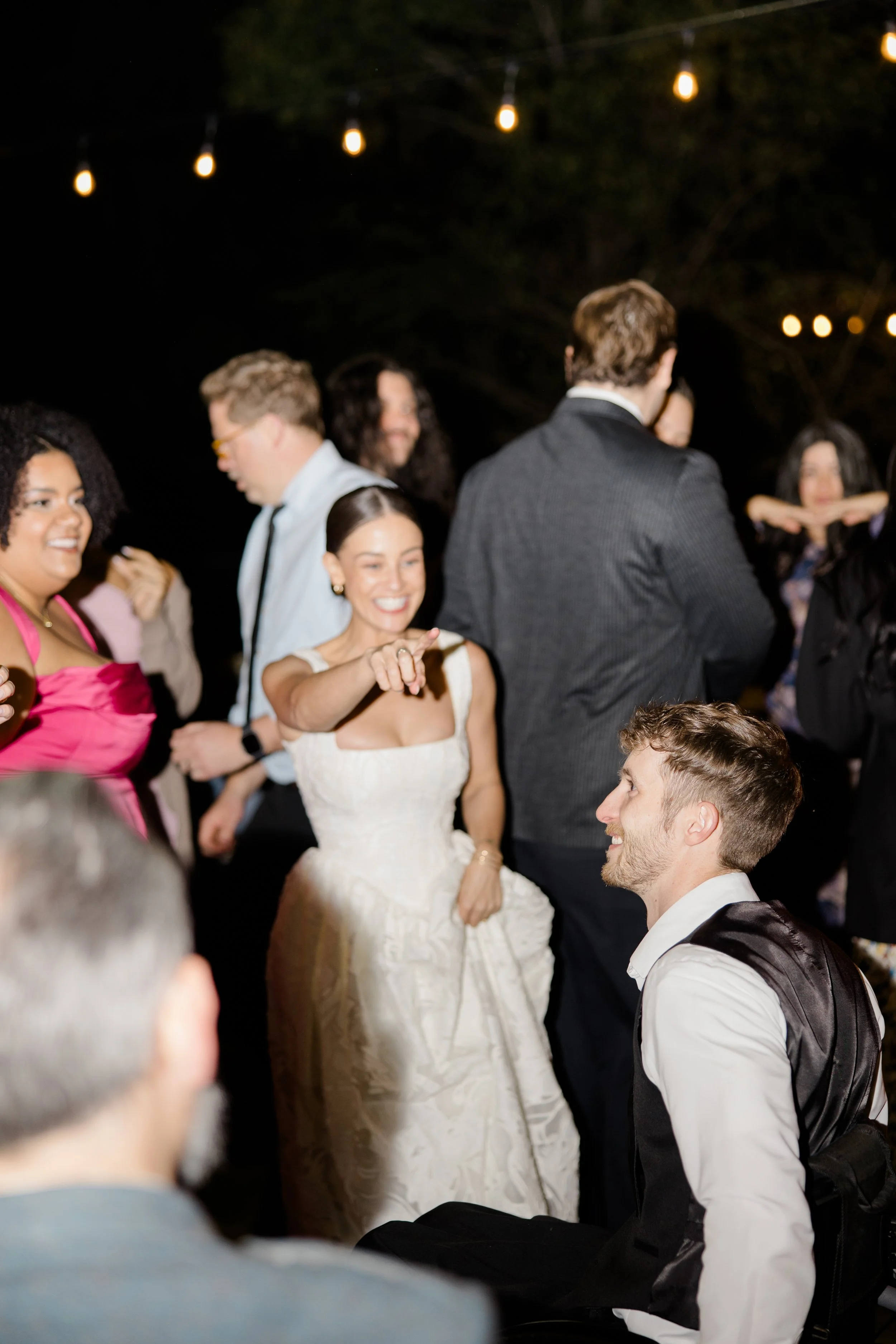 People dancing at an outdoor night event under string lights, including a woman in a white dress and a man in a vest, surrounded by smiling guests.