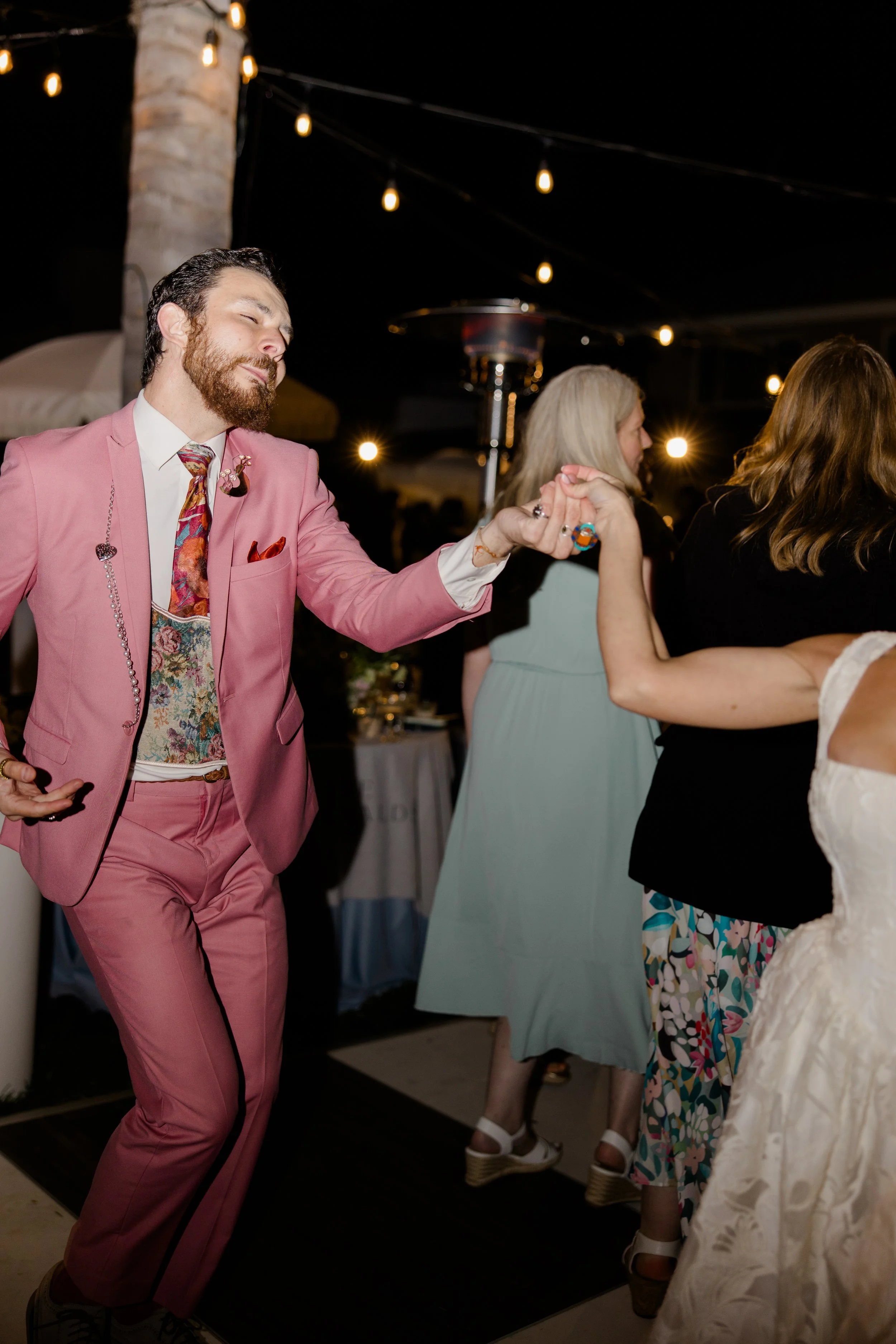 A man with a beard in a pink suit and floral tie is dancing with a woman at a night event with string lights overhead.