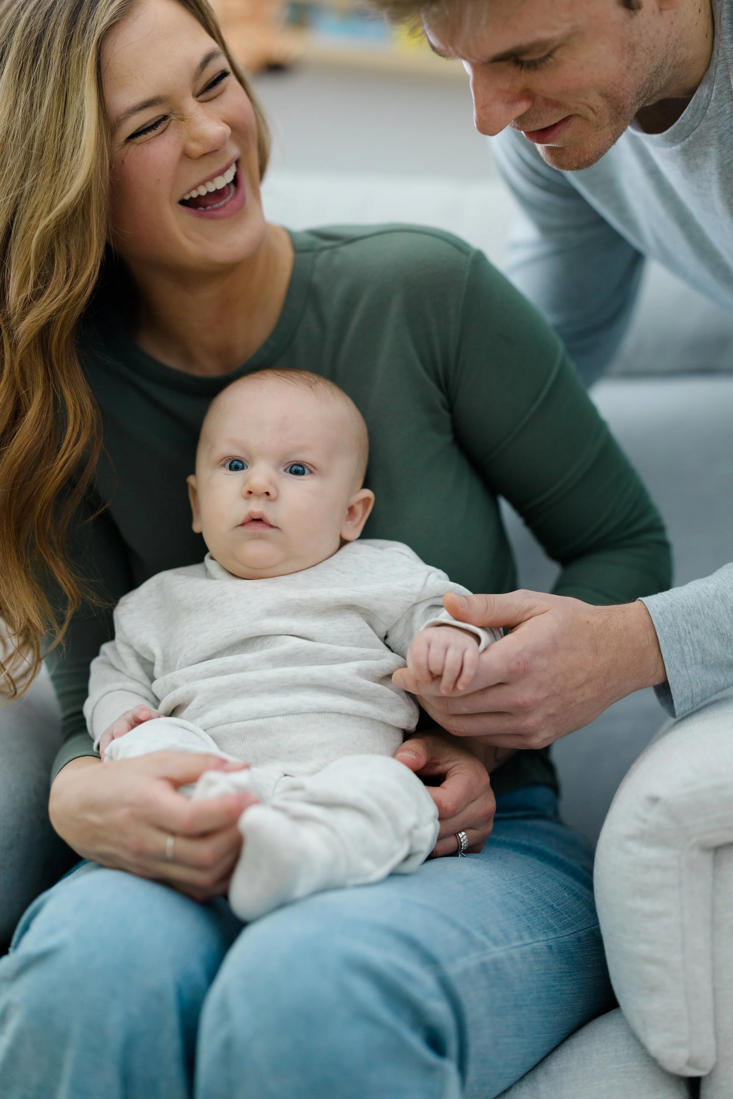 A woman and a man, possibly parents, hold a baby while sitting close together. The woman is smiling and appears to be laughing, and the baby has a neutral expression and blue eyes. The setting seems to be indoors, possibly in a home or a cozy environ