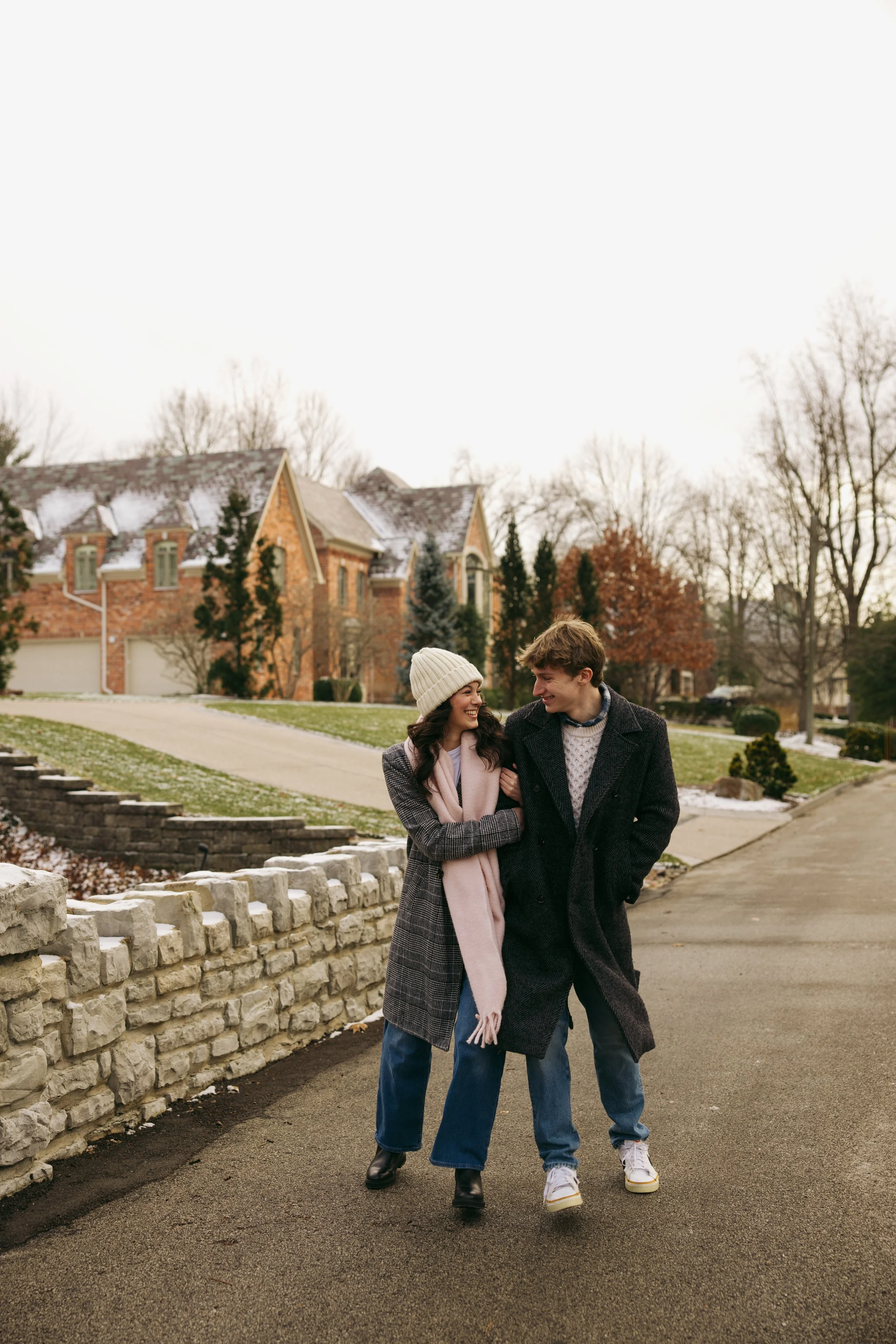 A young couple walking arm-in-arm on a sidewalk in front of a stone wall, with a large house and trees in the background, during winter.